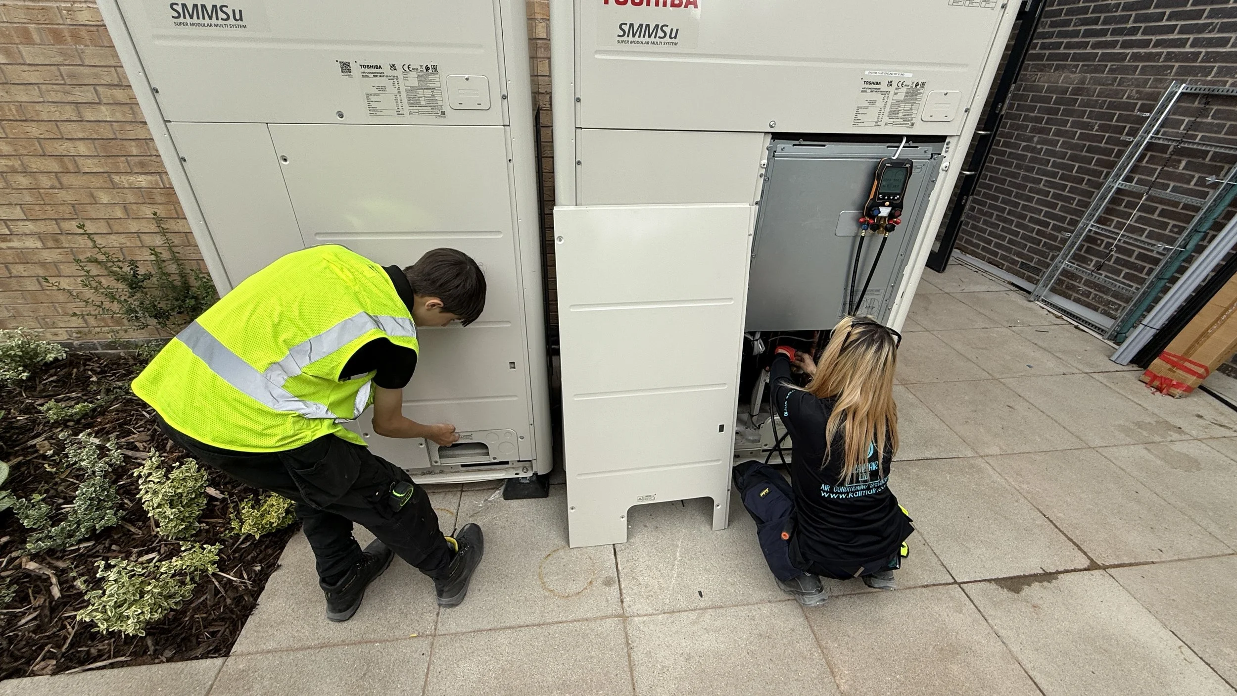 Two technicians wearing Kalmair Ltd uniform working on a large outdoor air conditioning unit on a sidewalk. One is kneeling and the other is crouching, inspecting and repairing the unit.
