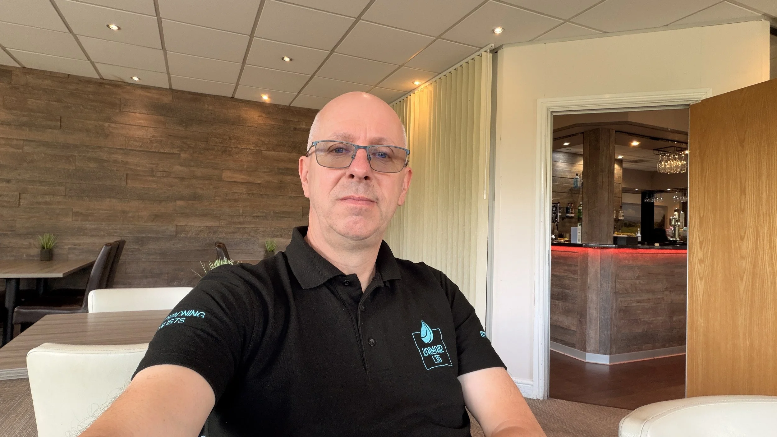 A man wearing glasses and a black collared shirt smartly dressed in uniform with a blue KALMAIR LTD  logo, sitting in a restaurant or cafe, taking a selfie. The background features wooden walls, a ceiling with recessed lighting