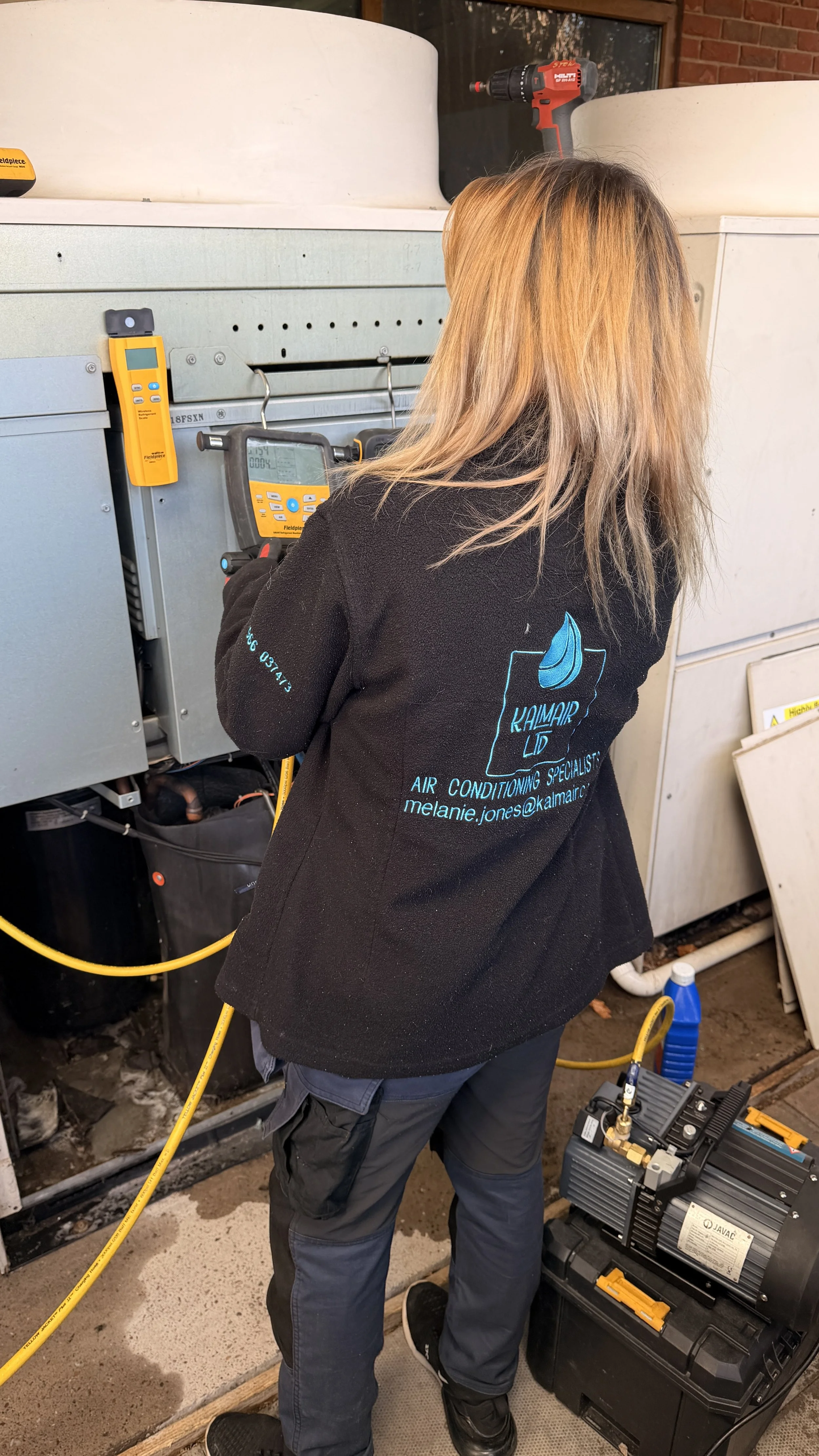 A person with blonde hair wearing a KALMAIR LTD standard uniform working on HVAC equipment, holding a digital meter, with a yellow tube and a black compressor nearby.