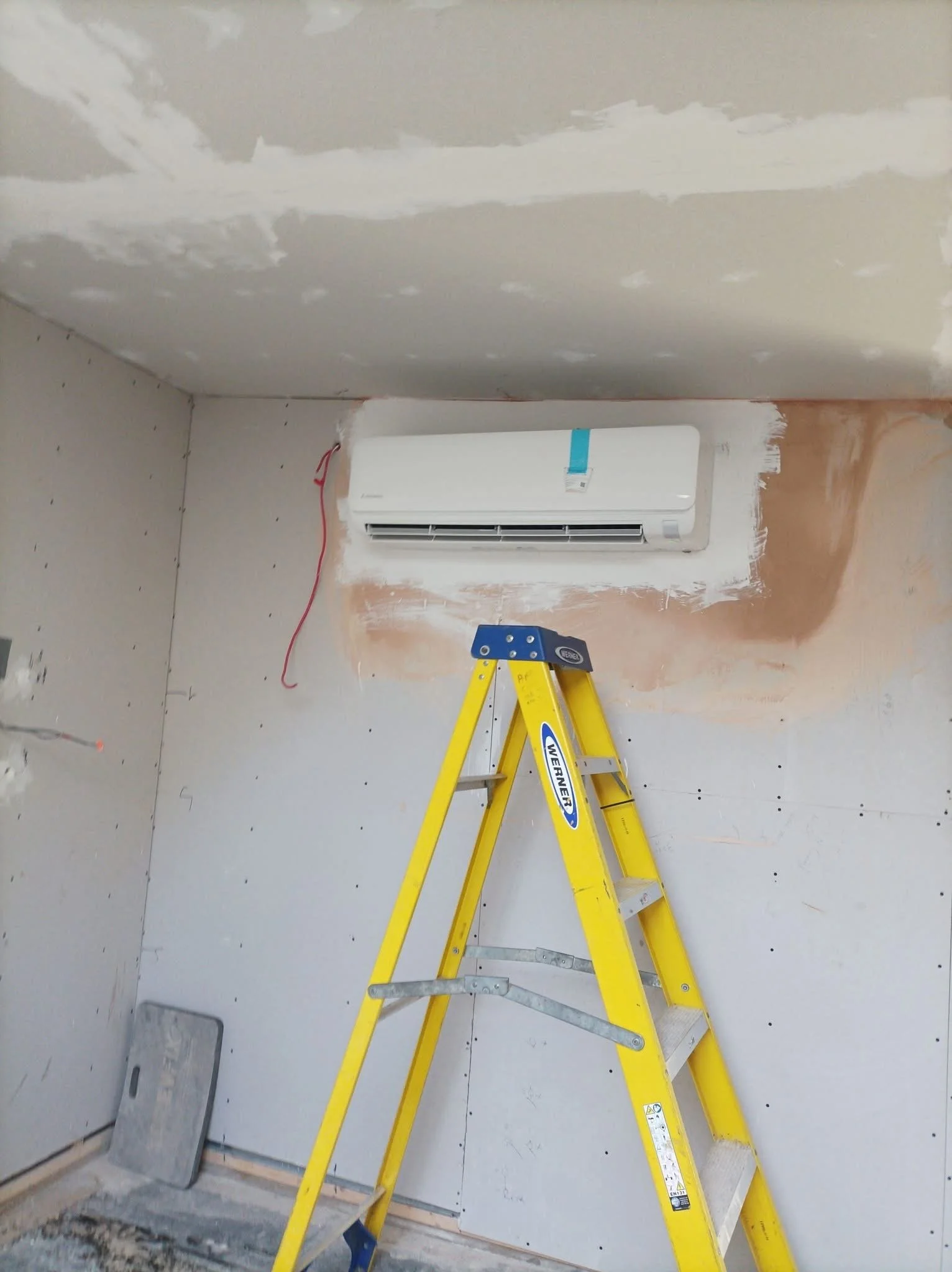 Yellow step ladder in a room under construction with drywall walls and ceiling, and a wall-mounted air conditioner unit.