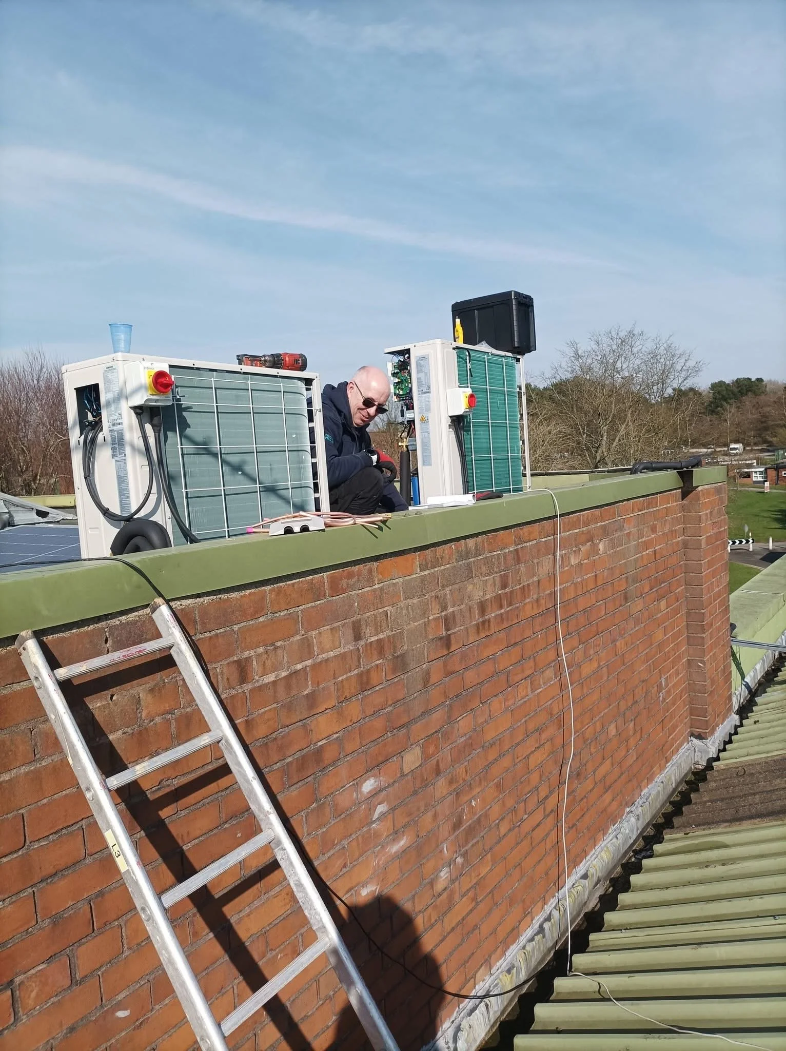 Man working on rooftop with Samsung HVAC units and solar panels, ladder leaning against brick wall, trees and blue sky in background.
