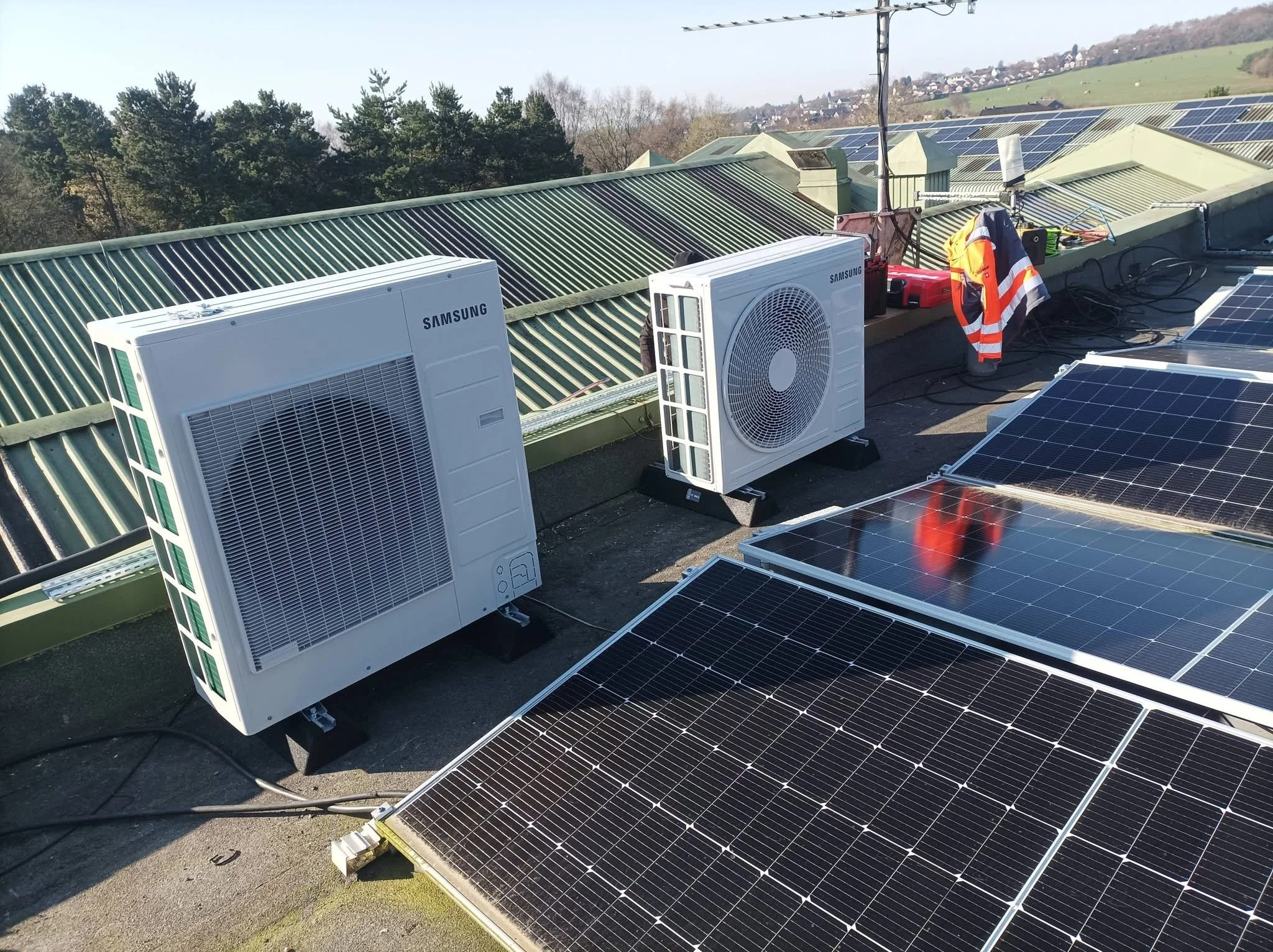 Solar panels and Samsung air conditioning units installed on a rooftop with trees and hills in the background.