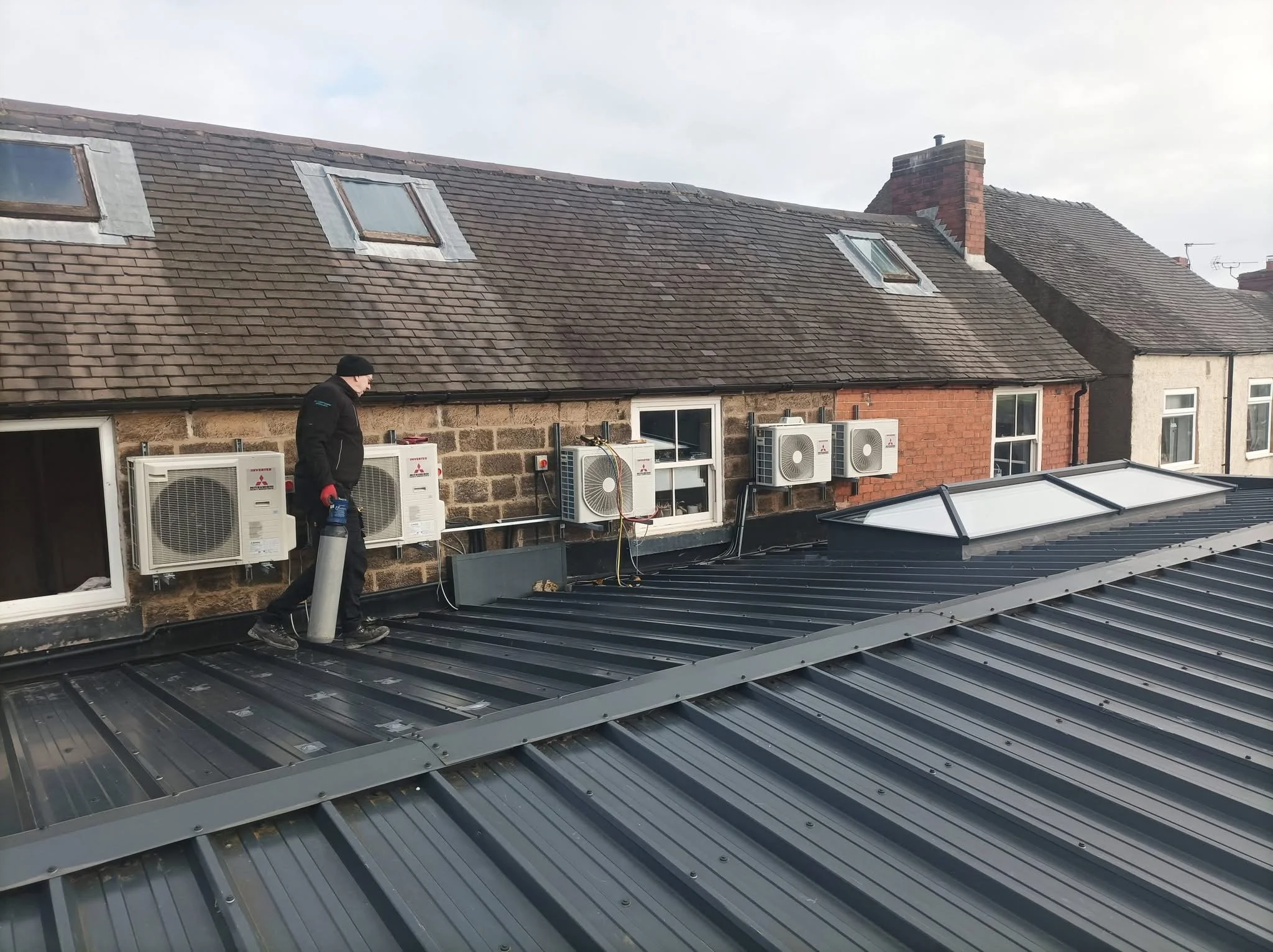 A person working on a rooftop with skylights, air conditioning units, and metal roofing, with neighboring houses and a chimney in the background.