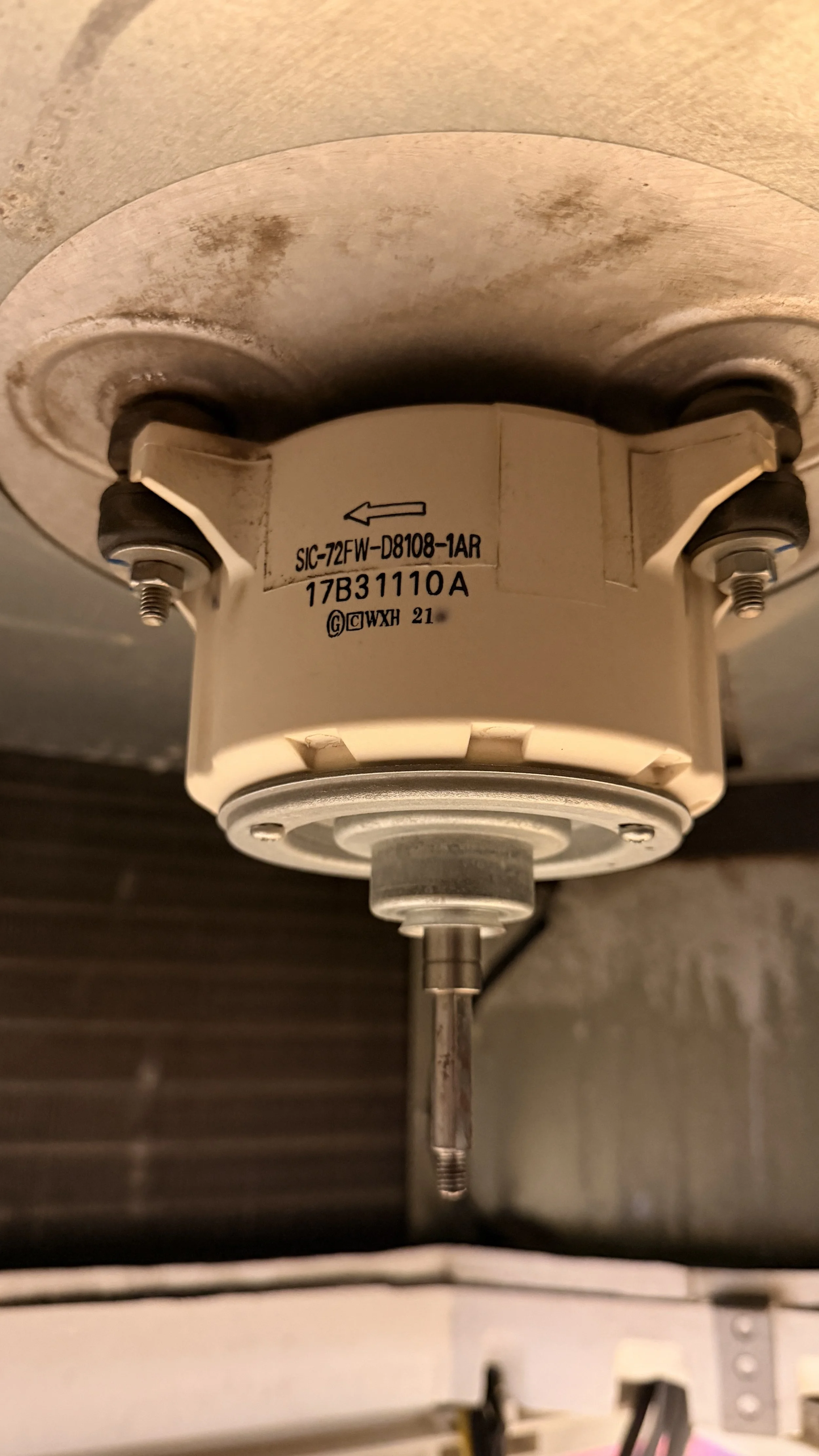 Close-up of an air conditioning fan motor with a white label containing black text and an arrow. The component appears aged or dirty with some grime or corrosion around it.