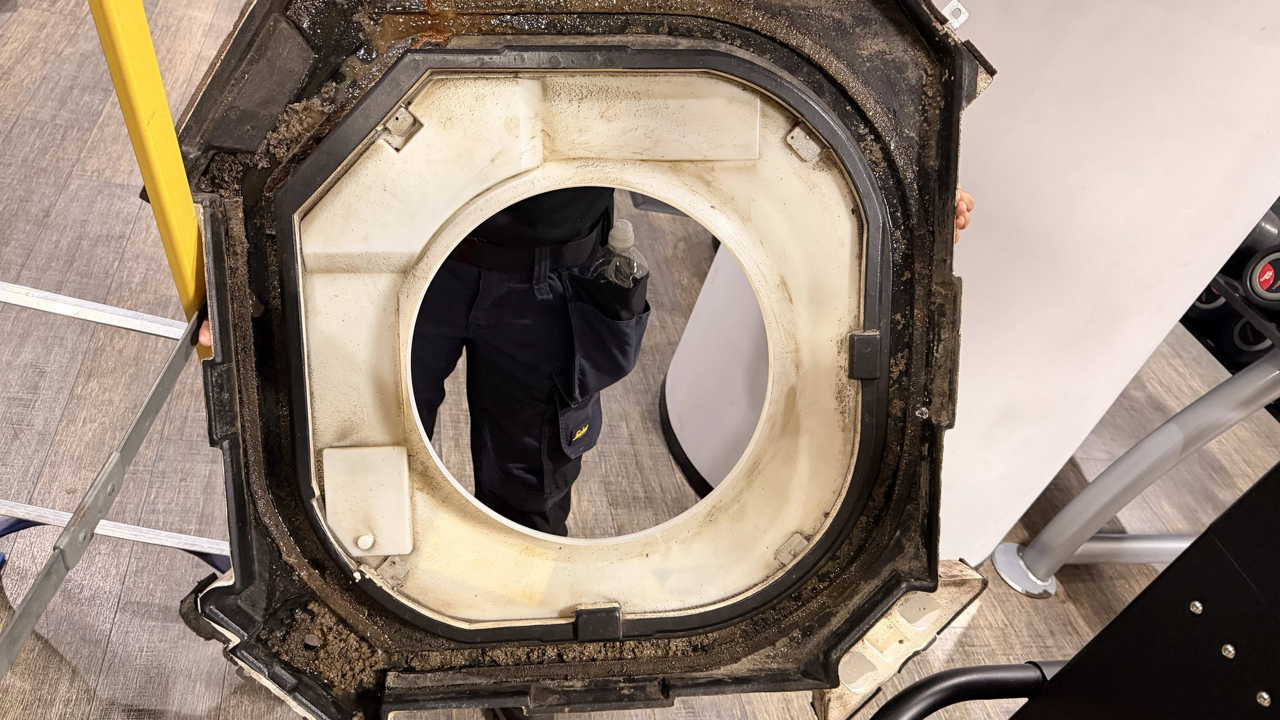 Close-up of an air conditioning cassette drip tray, showing dirt and grime around the edges, held by a person in black clothing. The device appears to be a large, circular mechanical or electronic component.