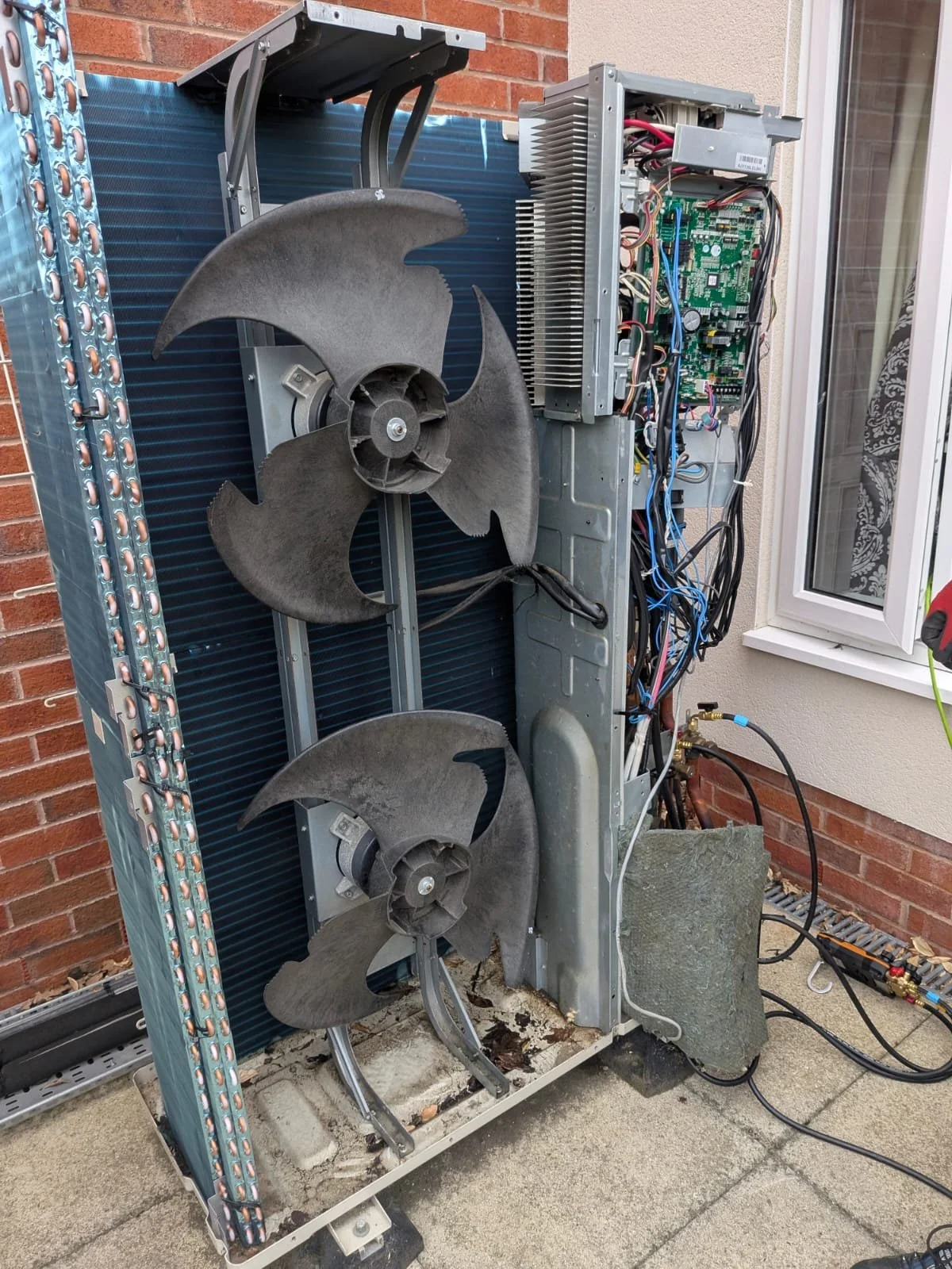 Open outdoor air conditioning unit with large fan blades, exposed circuit board, and wiring, located outside near a brick wall and window.