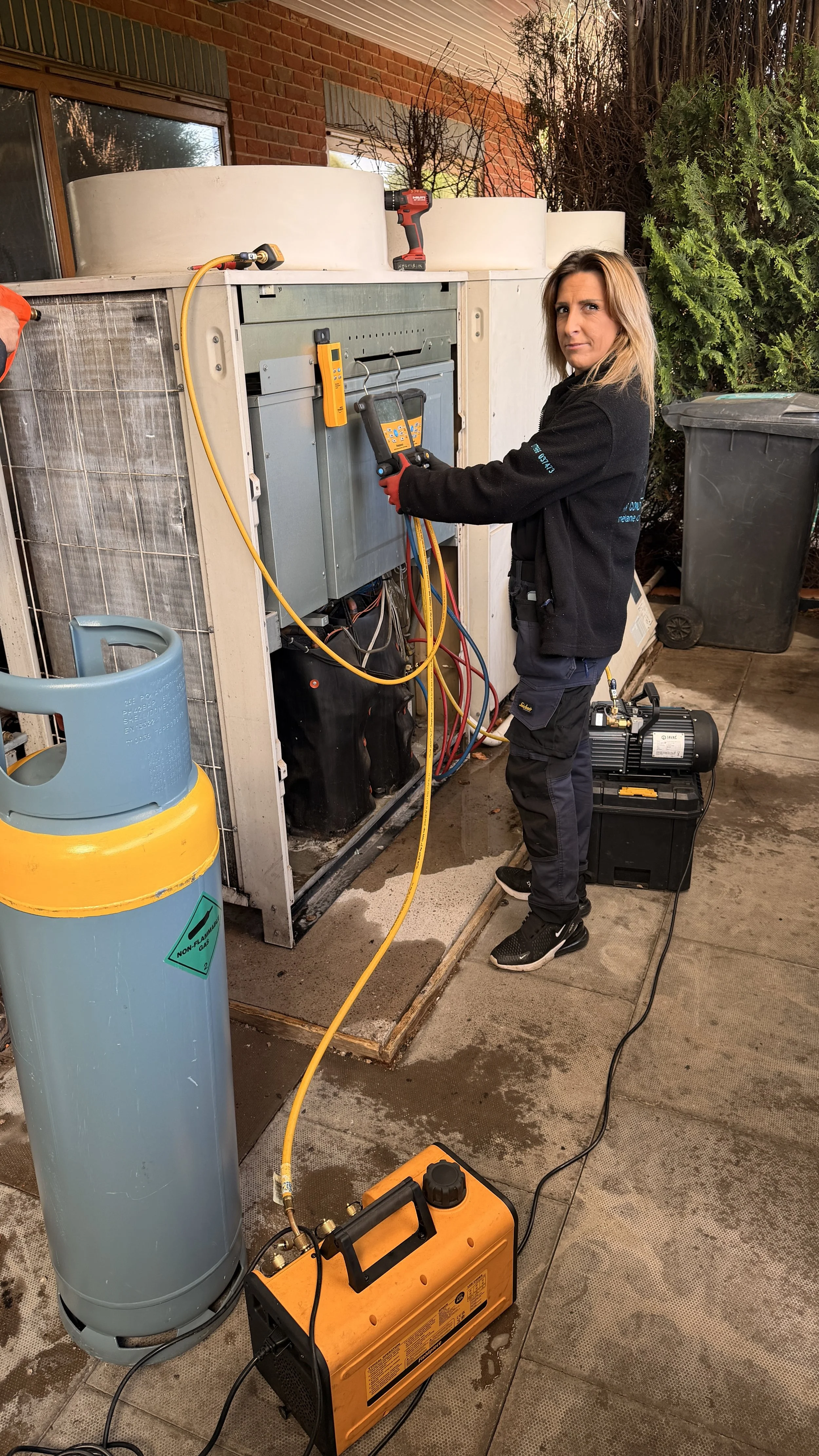 A technician wearing KALMAIR LTD standard uniform working on an outdoor central air conditioning unit with various tools and equipment, including a digital multimeter, vacuum pump, and refrigerant gauge.