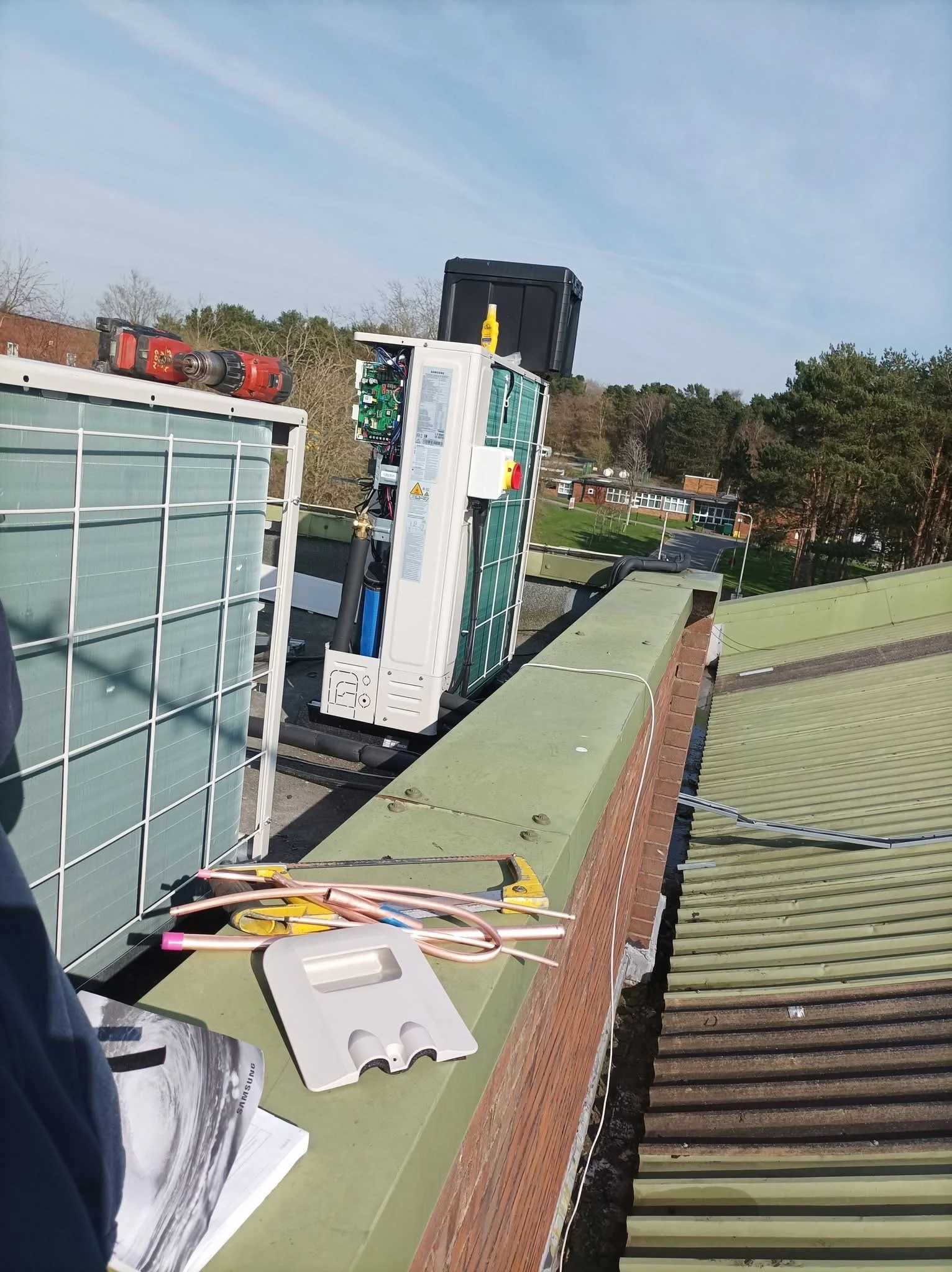 View of a rooftop with Samsung HVAC equipment, tools, and supplies, overlooking a suburban neighborhood with trees and houses, under a partly cloudy sky.
