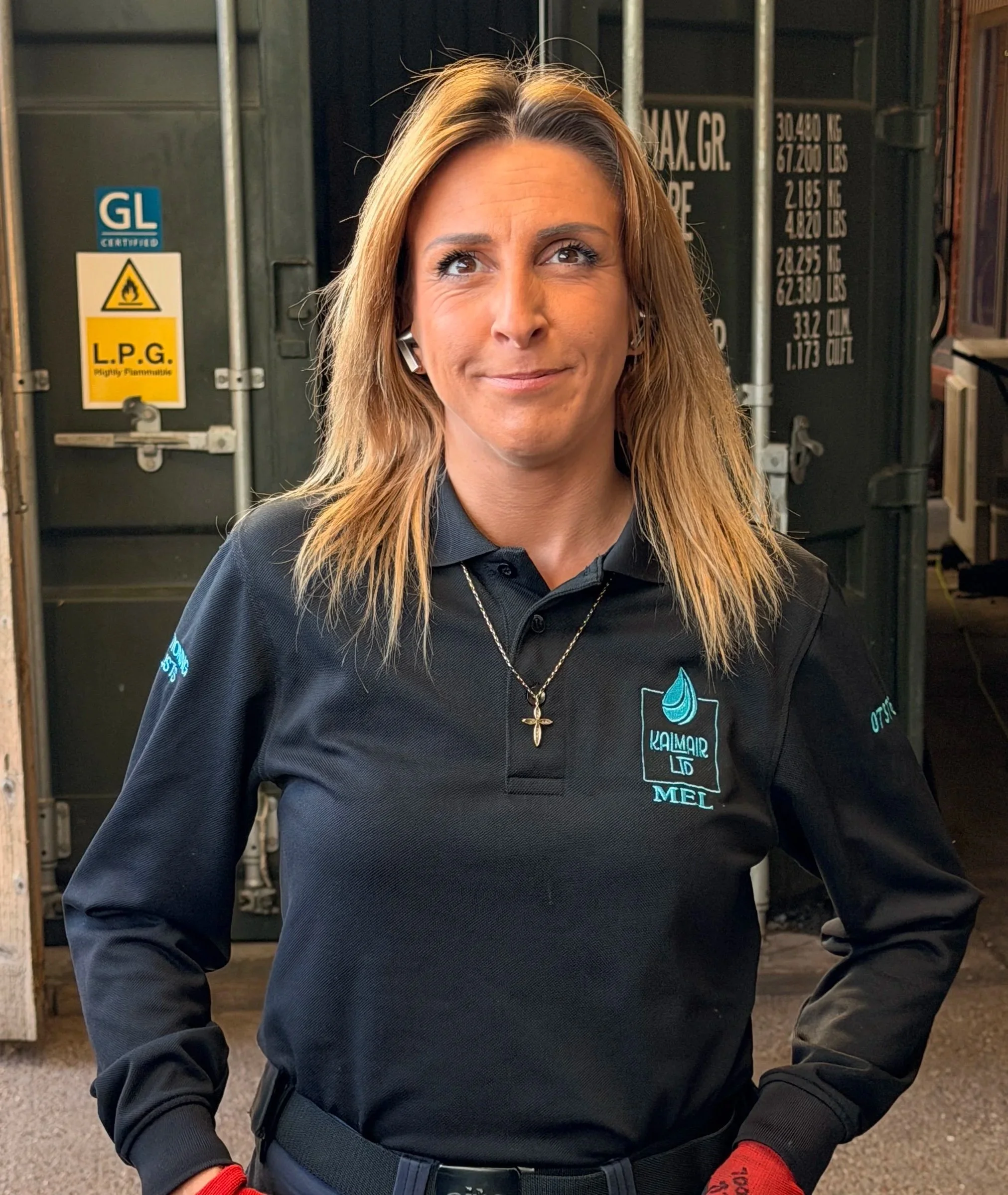 A woman wearing a black shirt with embroidered logos and text, a cross necklace, and red gloves, standing outside near a large industrial container with safety labels.