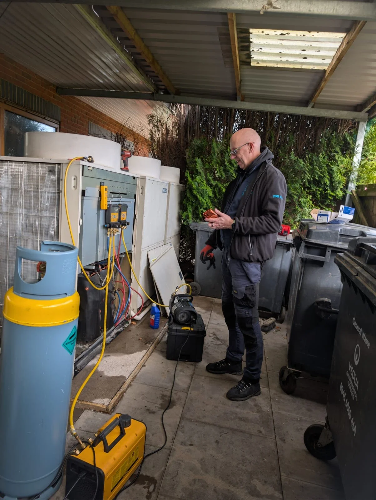 A man working on HVAC system outdoors wearing a KALMAIR LTD standard uniform, surrounded by tools and equipment, with a yellow refrigerant tank nearby and trash bins in the background.