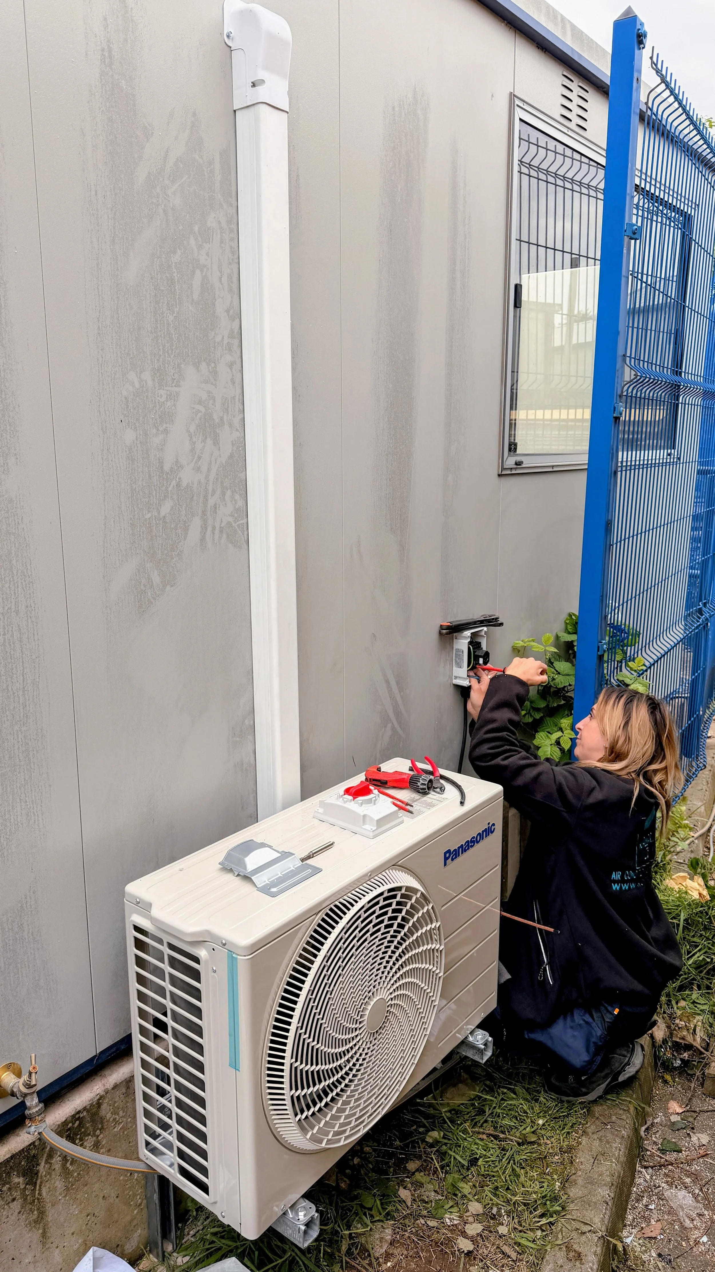A woman wearing KALMAIR LTD standard uniform is kneeling outdoors working on an air conditioning unit mounted on an exterior wall, with tools on a nearby white box and a blue security fence in the background.