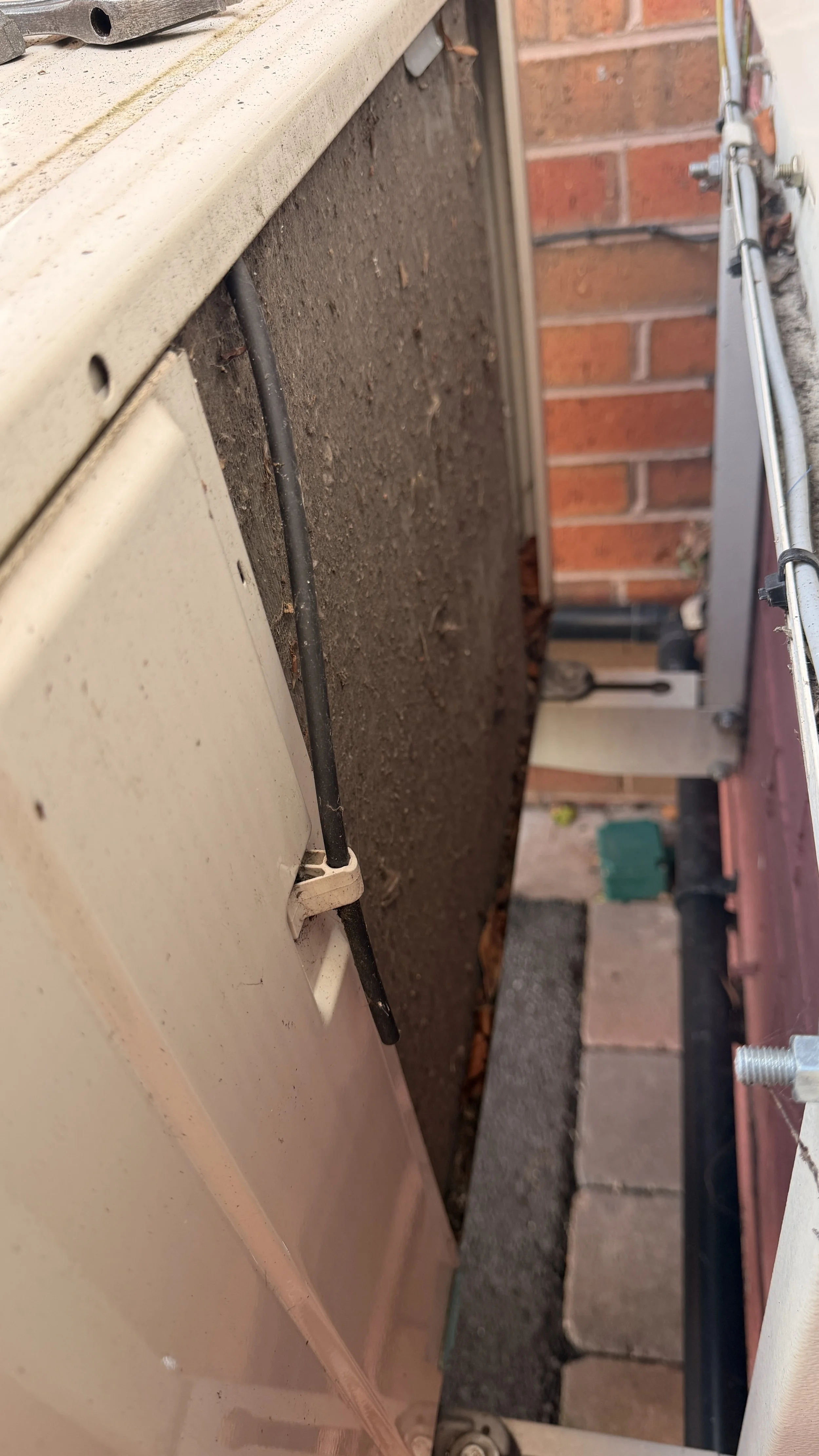 Close-up of a beige outdoor air conditioning unit or equipment with black hoses and a brick wall in the background.