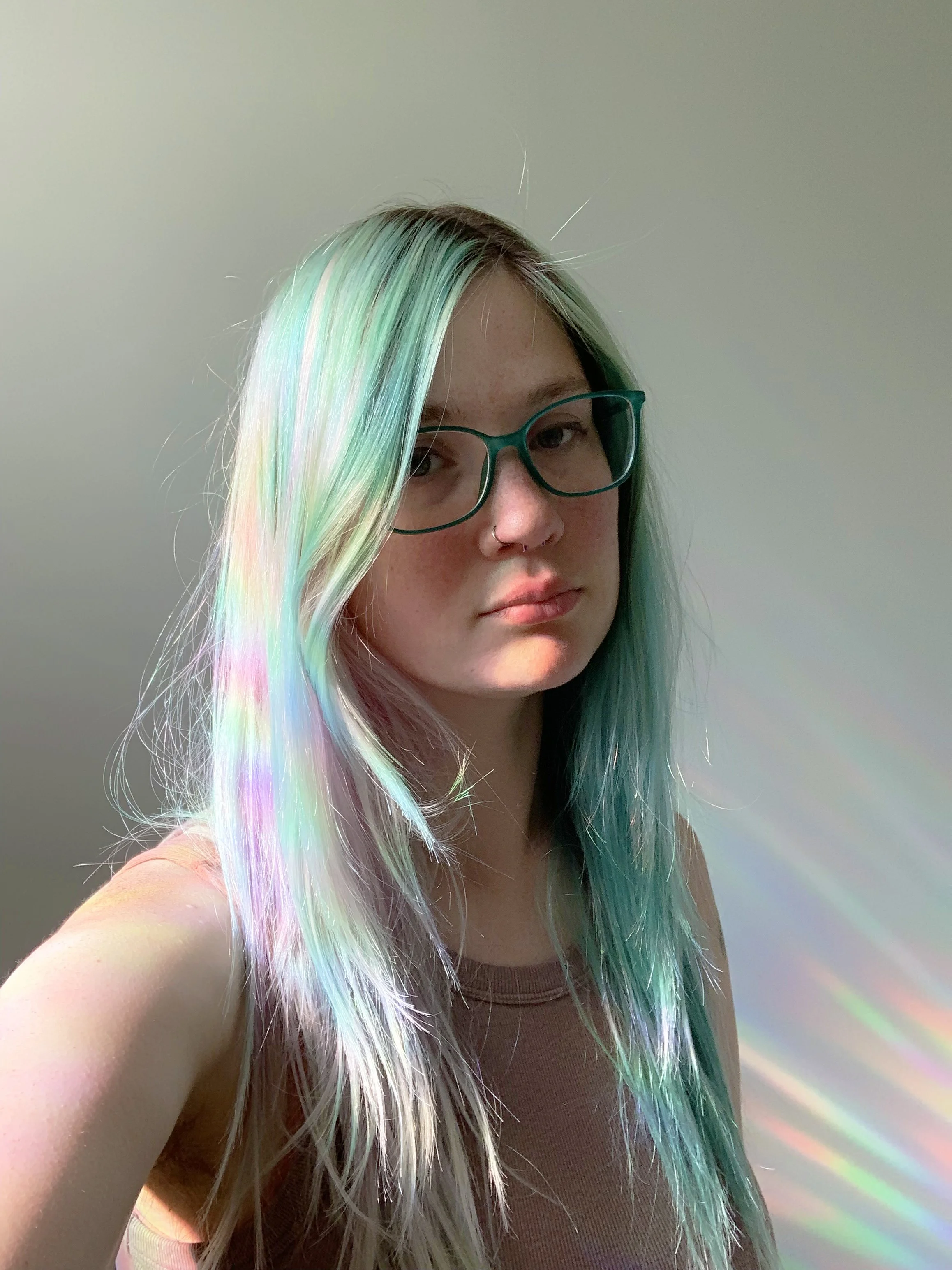 Headshot of a feminine presenting person with long blue hair, glasses and nose piercings standing in front of a white wall. There is a rainbow pattern from a suncatcher cast onto the left side their hair and on the wall behind them on the right.