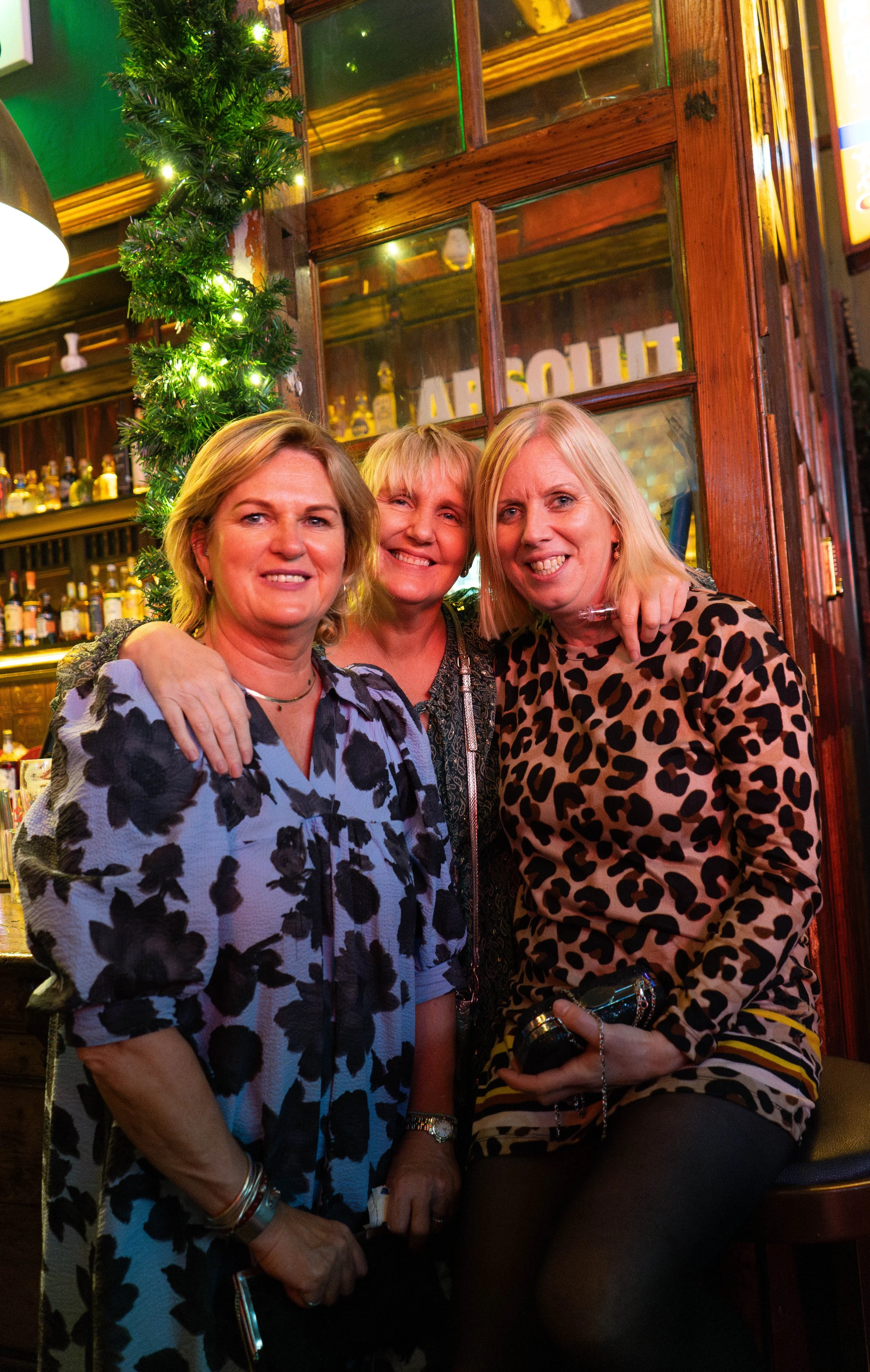 Three women smiling and posing together inside a bar decorated for the holidays, with a Christmas garland and a wooden bar in the background.