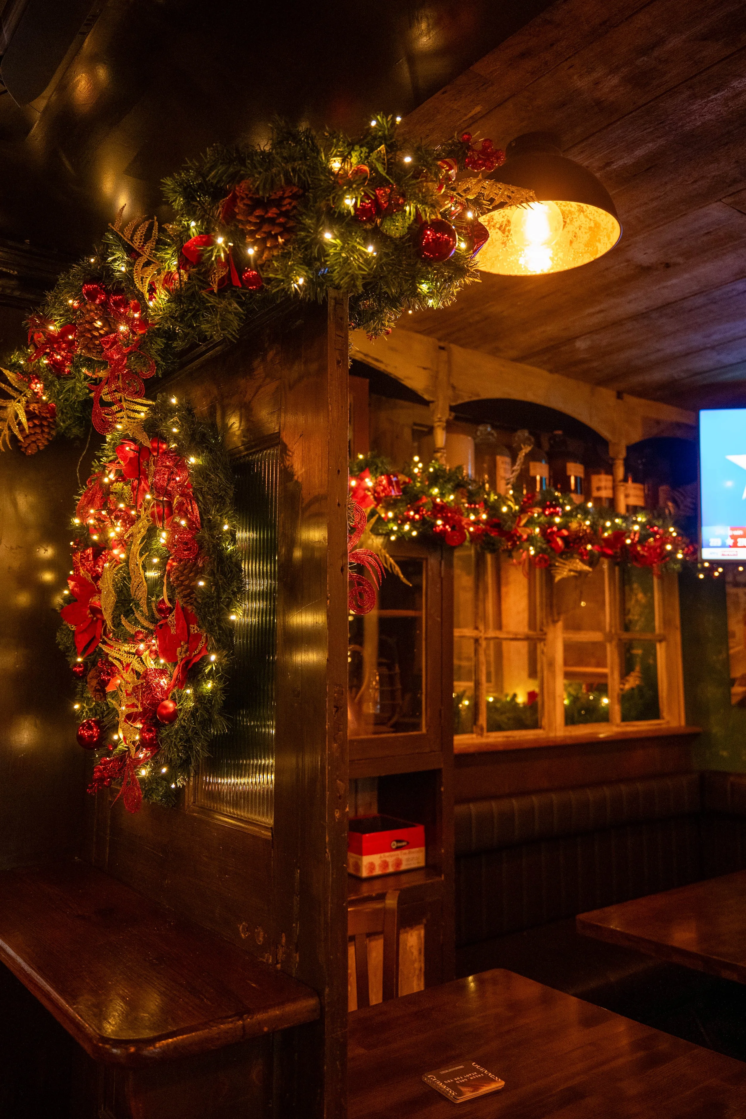 Festively decorated interior featuring a corner with Christmas garland, red ribbons, ornaments, pinecones, and string lights, warm lighting, wooden furniture, and cozy window views.