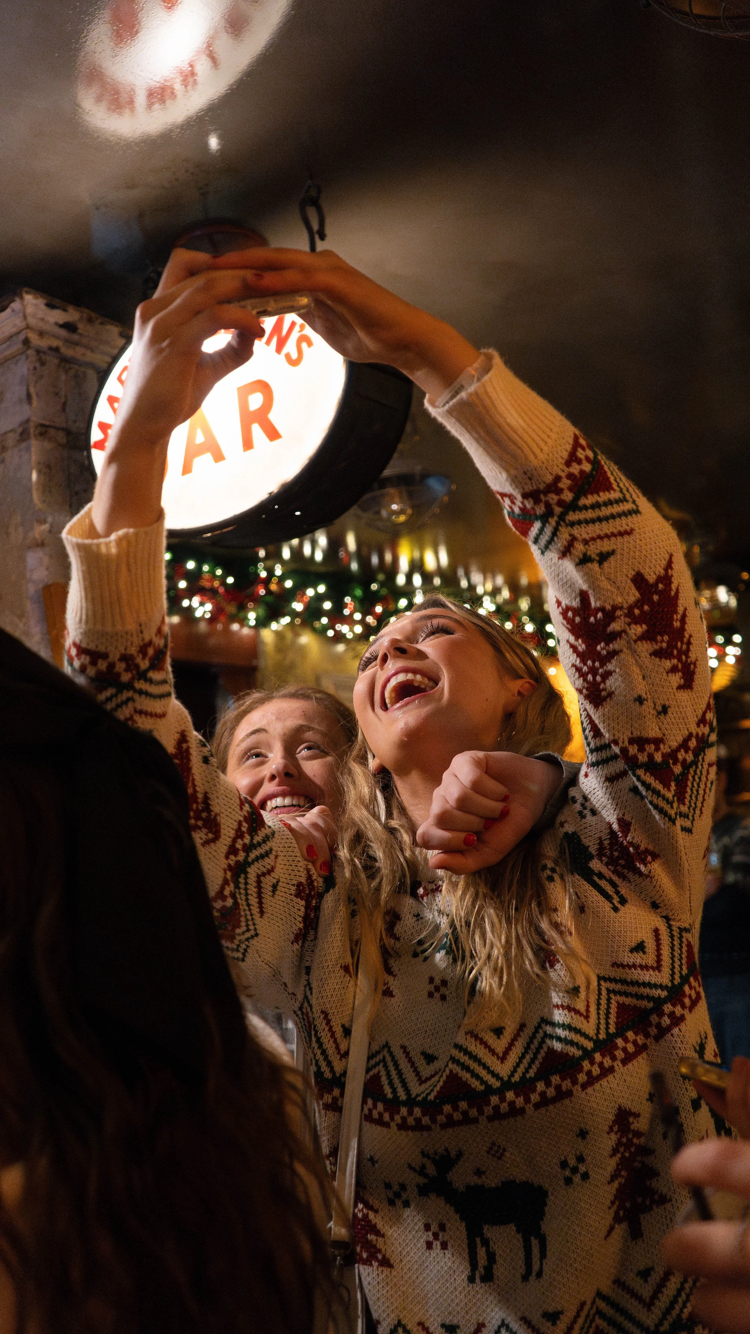Two girls wearing Christmas sweaters taking a selfie in a festive decorated bar with a sign overhead