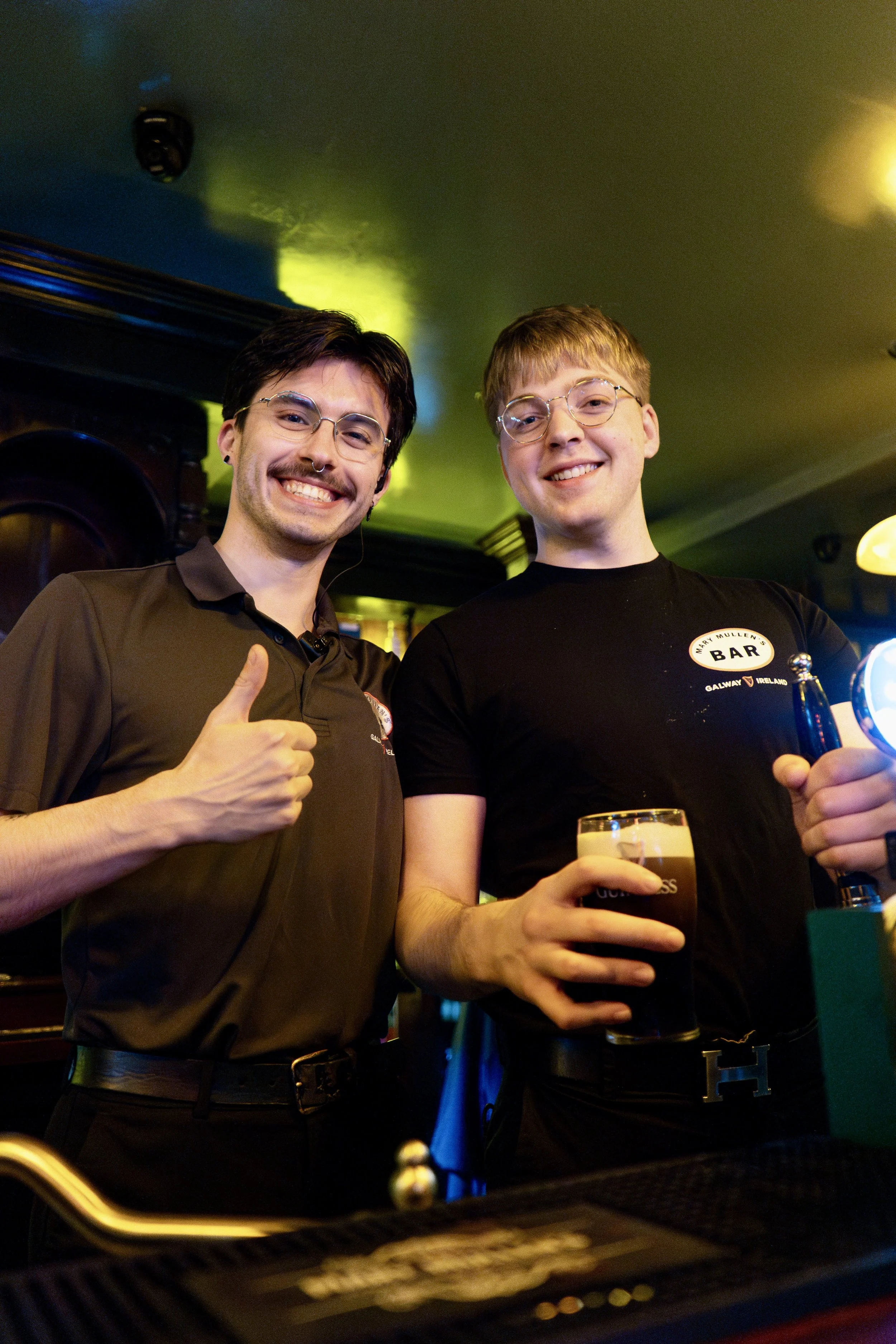 Two young men standing at a bar, smiling at the camera. One is giving a thumbs-up, the other holding a pint of dark beer. The bar has colorful lighting.
