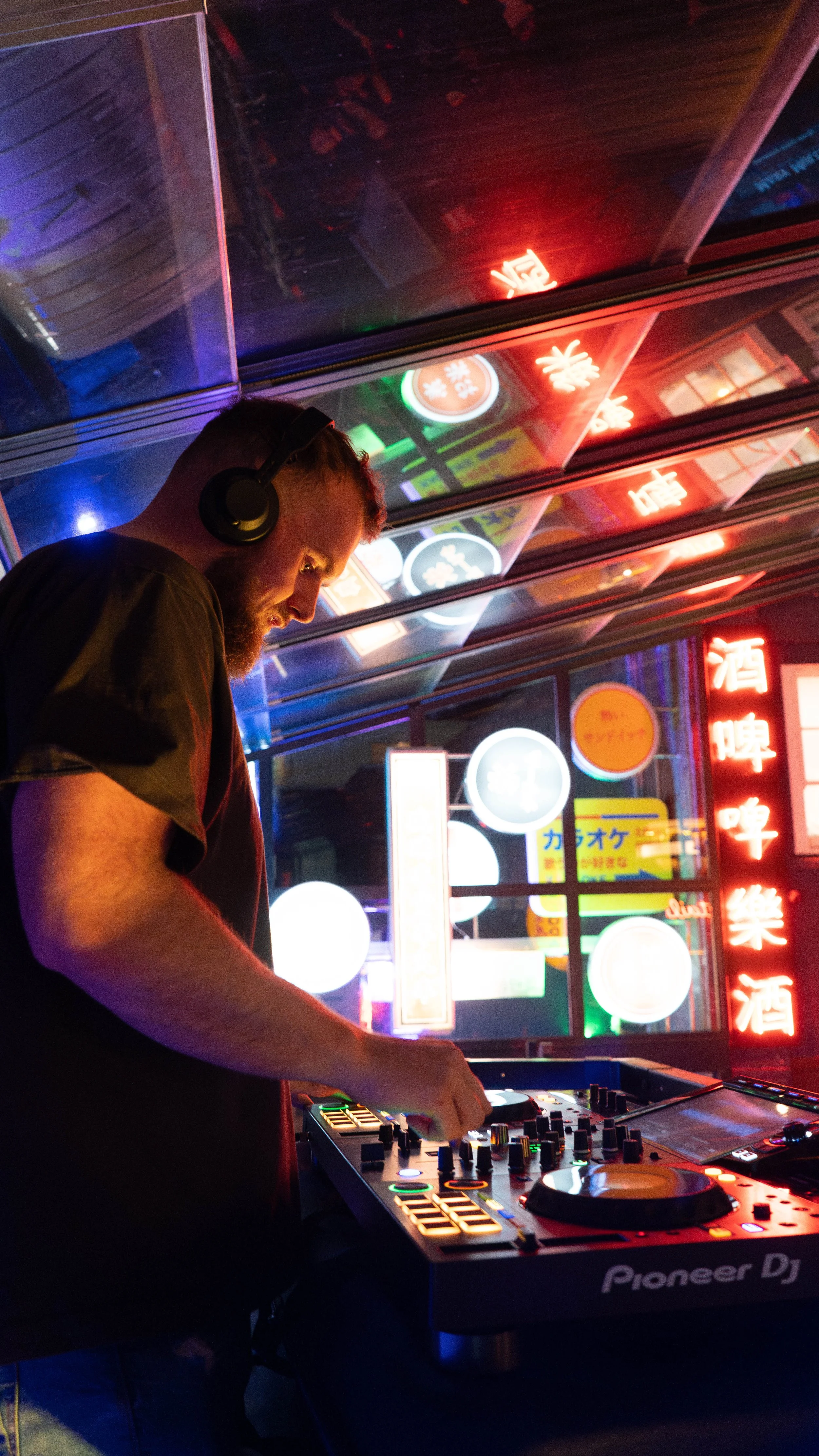 A DJ wearing headphones performs at a nightclub with neon signs and colorful lights visible through the window behind him.