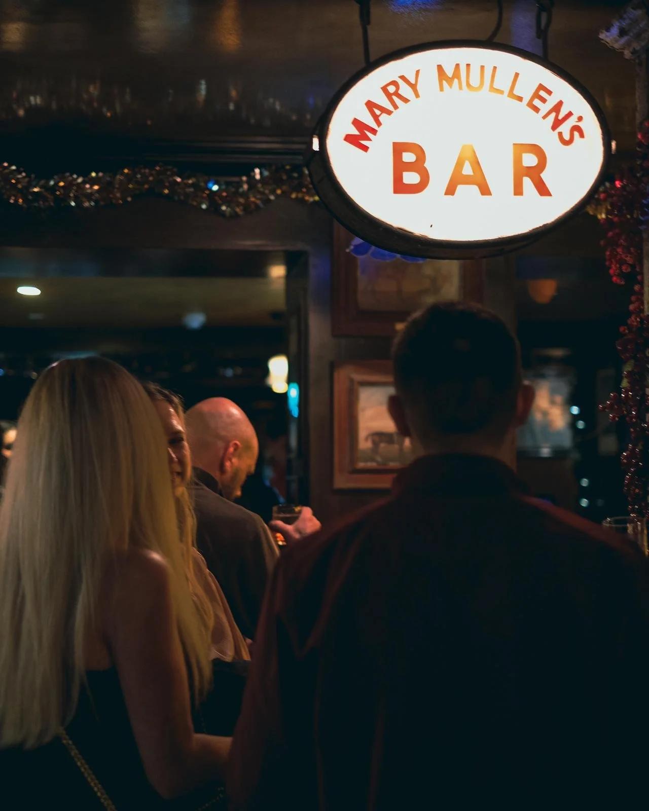 People inside a bar with a sign that reads 'Mary Mullen's Bar' hanging overhead.