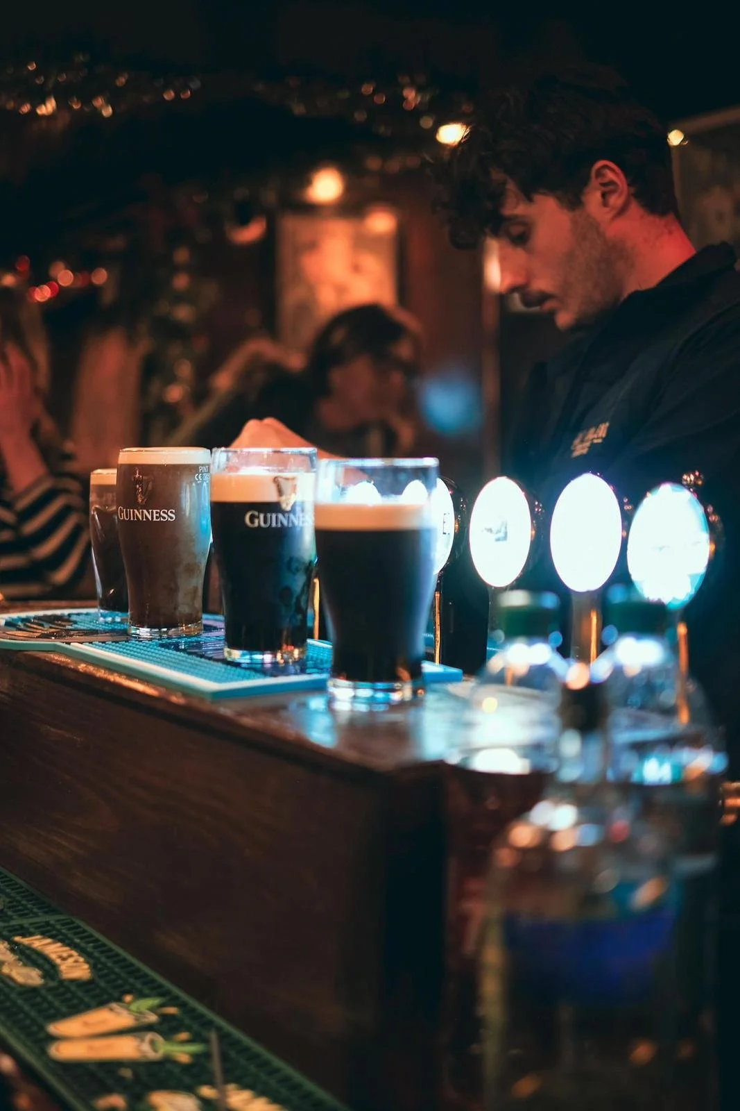 Several pints of Guinness beer on a bar counter in a dimly lit pub, with a bartender in the background.