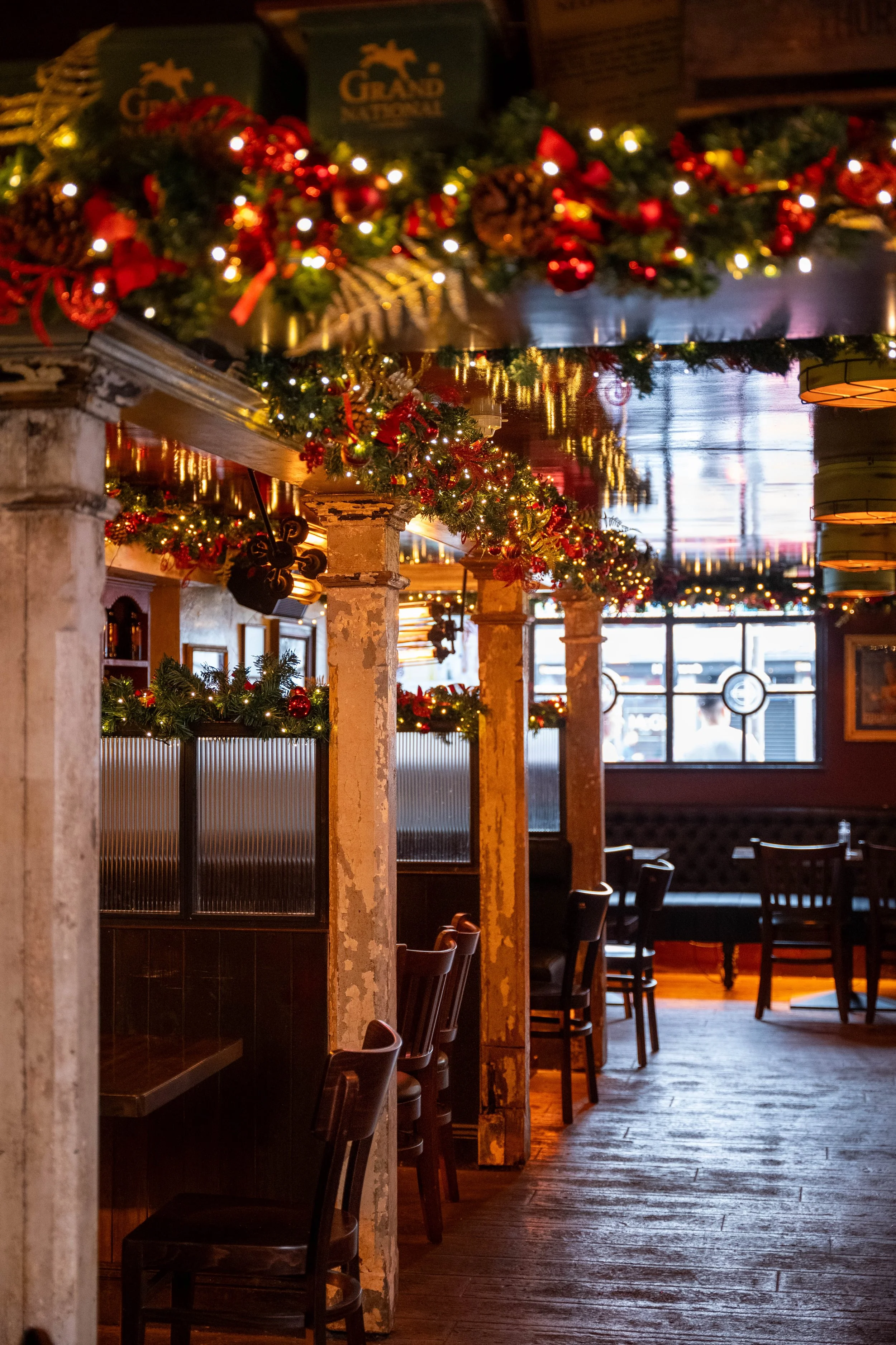 Interior of a restaurant decorated for Christmas with garlands, red ornaments, and string lights hanging from the ceiling and pillars, with empty chairs and tables.
