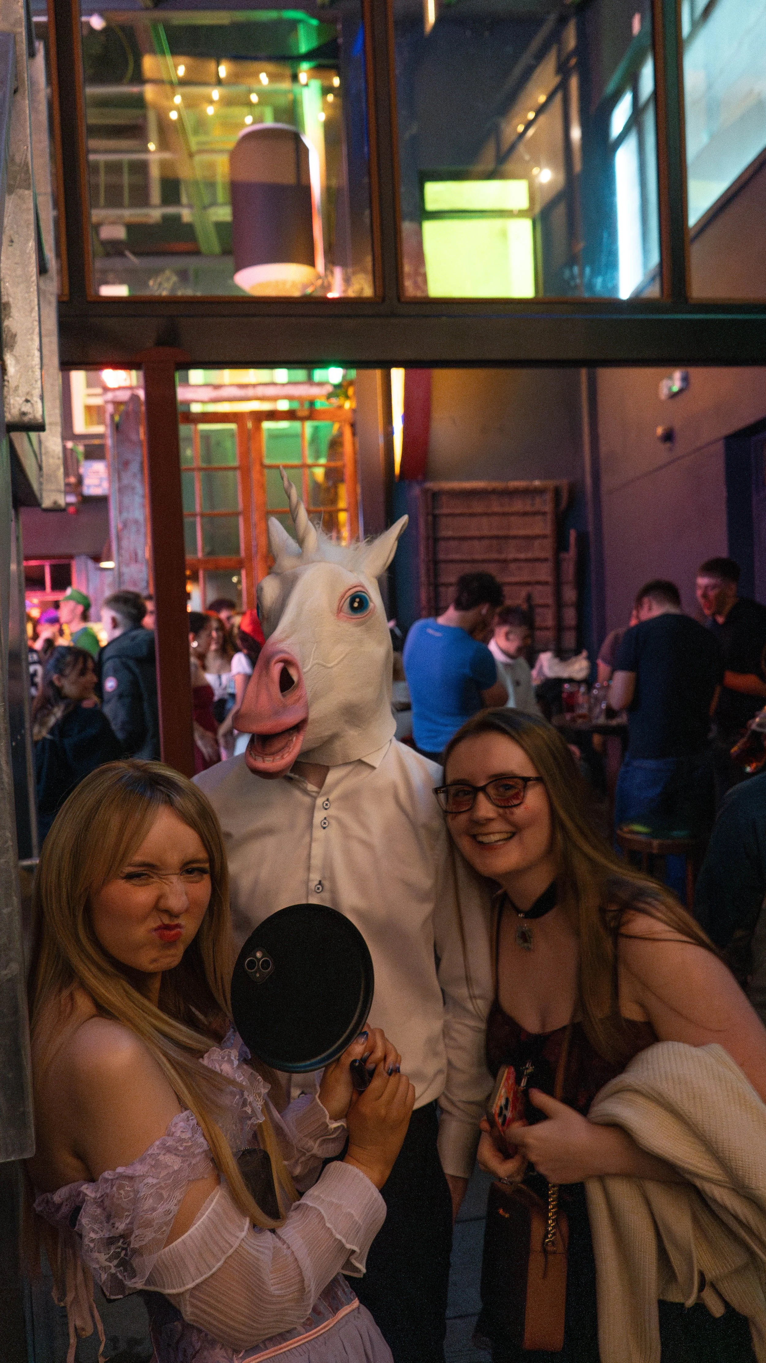 A group of friends at a lively party, with a person wearing a realistic unicorn mask, two women smiling and making playful expressions, one holding a frying pan, inside a vibrant, dimly-lit venue with colorful neon lights and a crowd in the backgroun