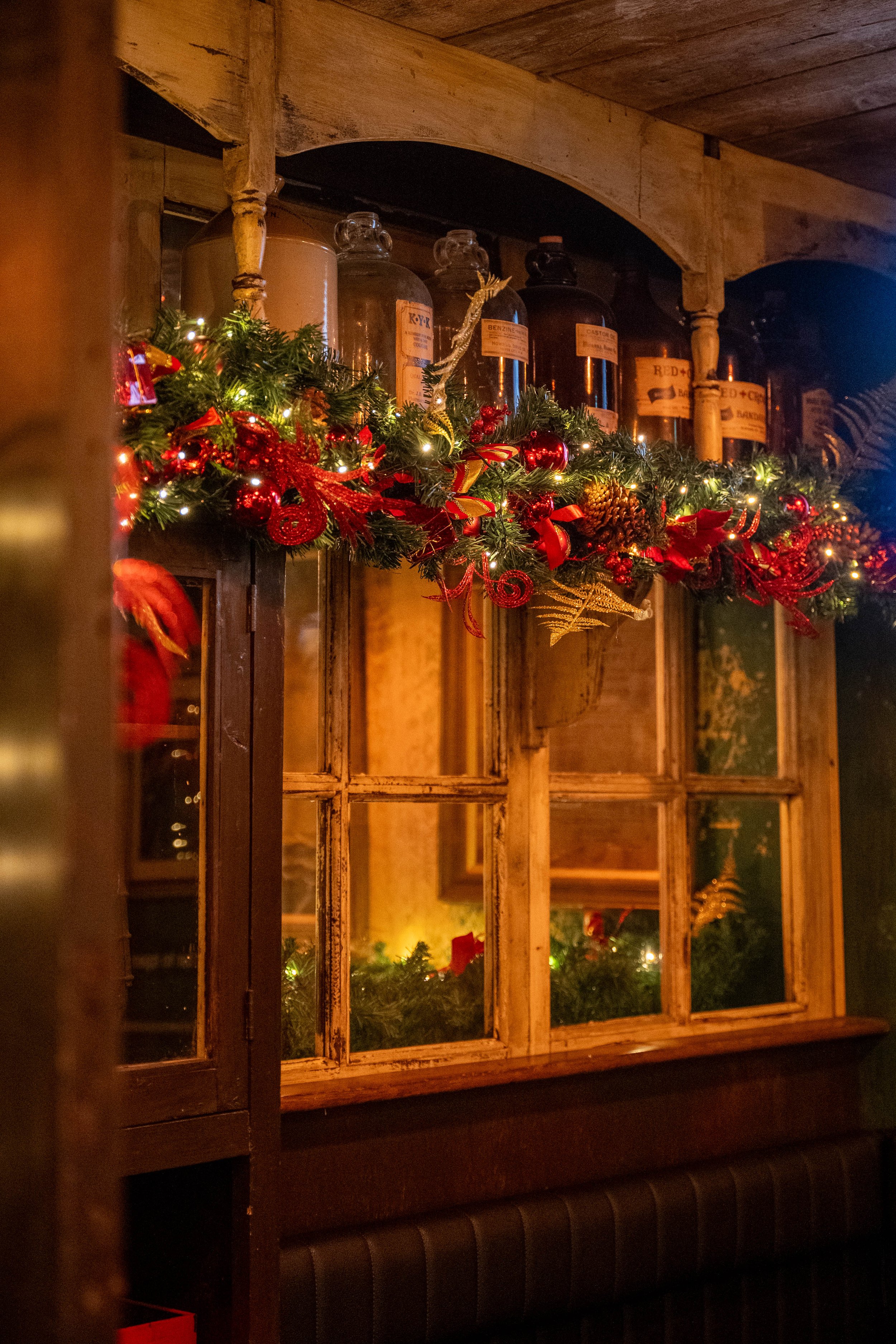 Decorative Christmas garland with red ribbons and pinecones hanging above a wooden window with bottles of alcohol on a shelf