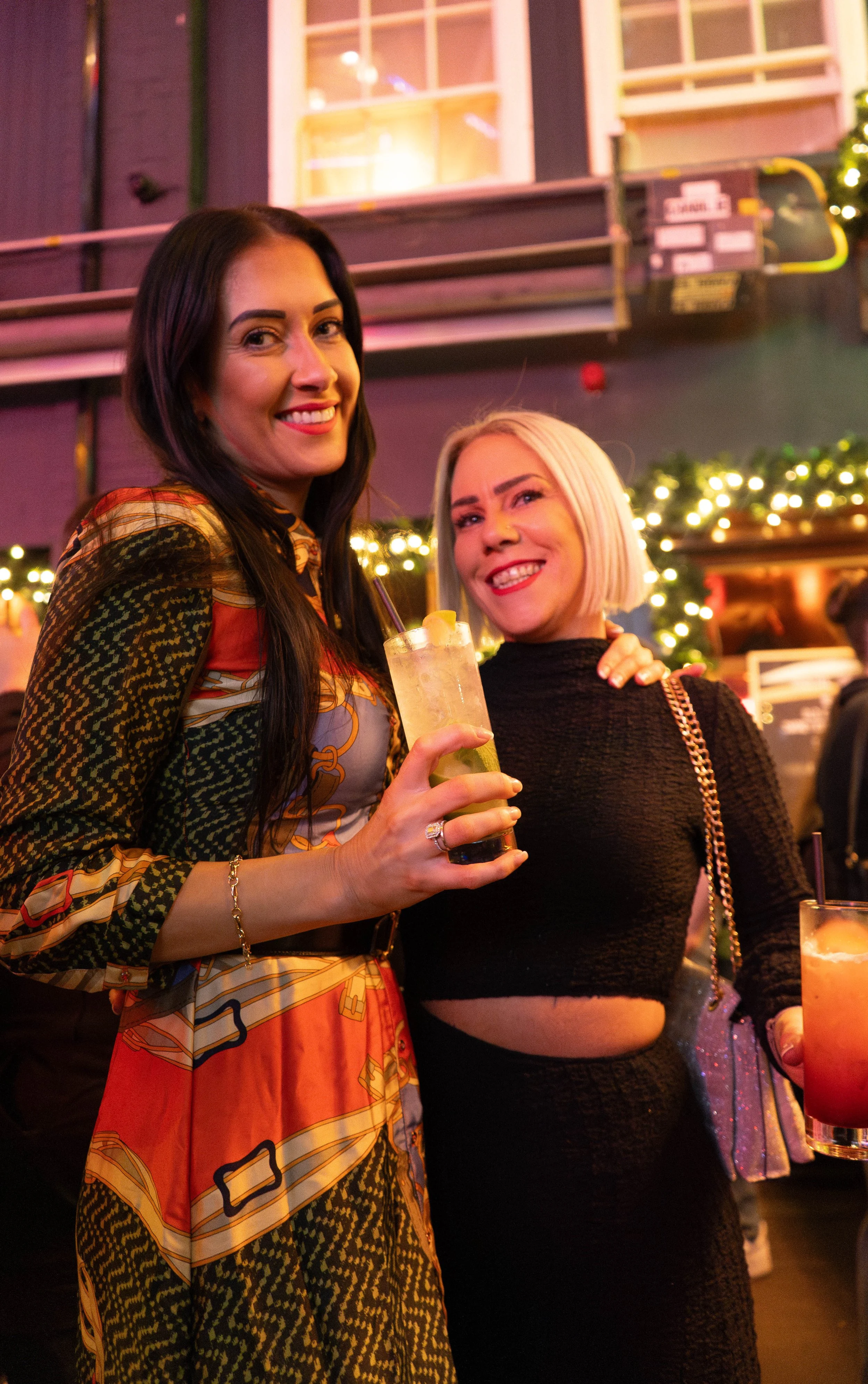 Two women celebrating at a bar during winter holiday season, holding drinks and smiling for the camera, with festive Christmas lights and decorations in the background.