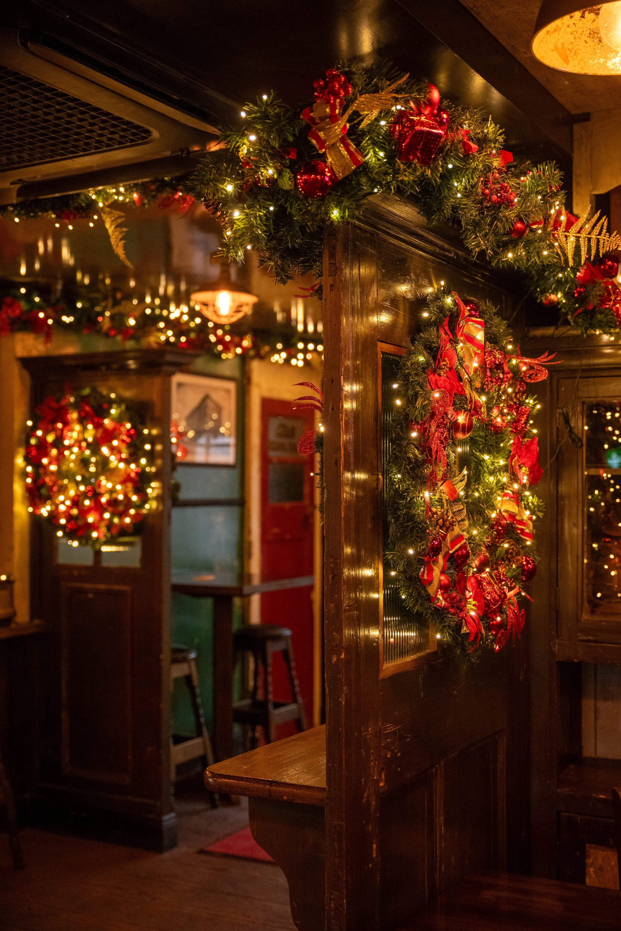 A cozy restaurant decorated for Christmas with wreaths and garlands of greenery, red ornaments, and ribbons, warm lighting, and wooden furniture.