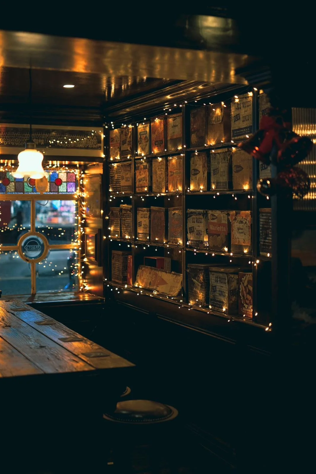 Cozy cafe interior with wooden counter, warm string lights, and bookshelf filled with colorful boxes or books, illuminated by soft warm lighting.