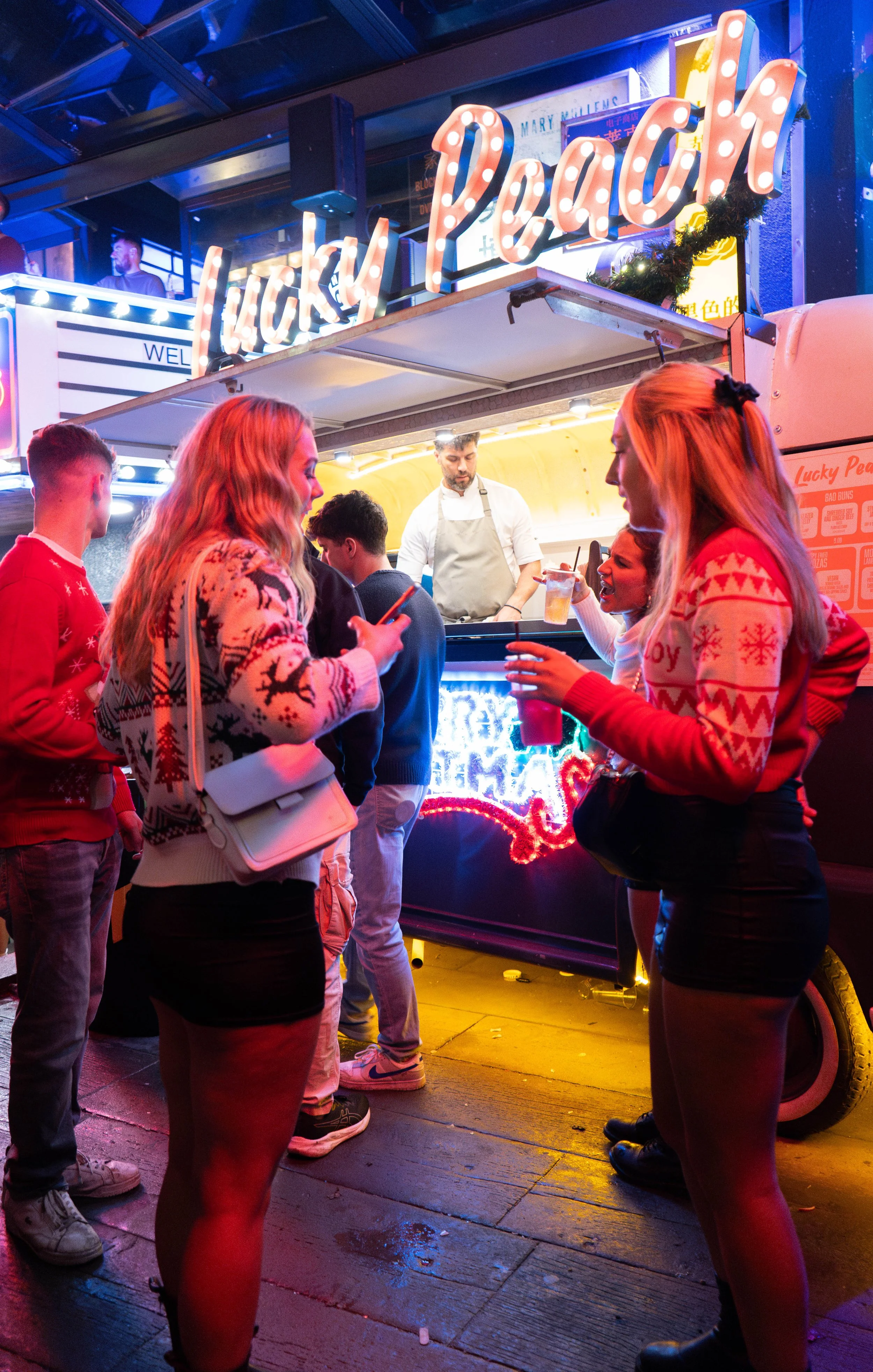 People standing at a night market stall called Lucky Peach, which has bright neon lights and a sign saying 'Lucky Peach'. A man in an apron is preparing food behind the stall, while women in festive sweaters are ordering beverages. The scene is livel