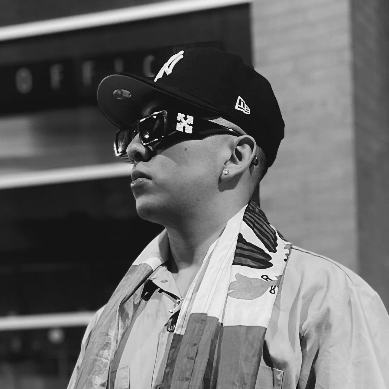 A young man wearing sunglasses, a black cap, earrings, and a patterned shirt, standing in profile against an urban background in black and white.