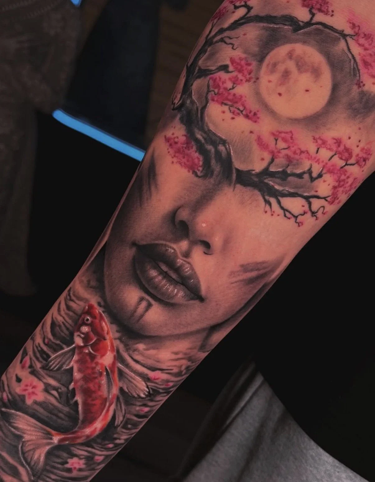 A tattoo featuring a woman's face with closed eyes, full lips, and a serene expression. Above her head, a tree with pink blossoms and a full moon in a night sky. A red koi fish is depicted below her face in water, with cherry blossoms around it.