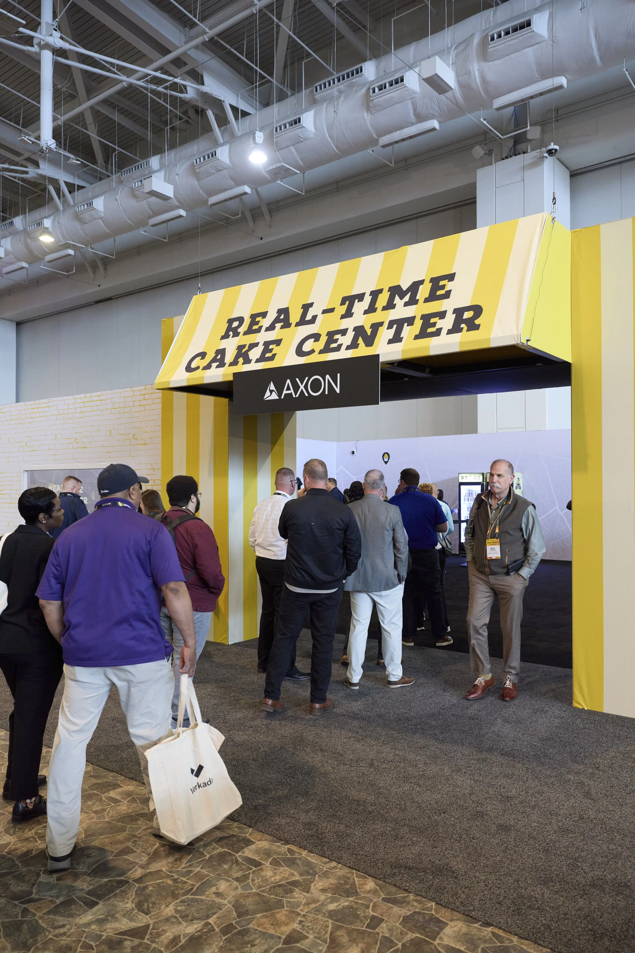 People gathered at a booth with yellow and black signage reading 'Real-Time Cake Center' at a conference or trade show.