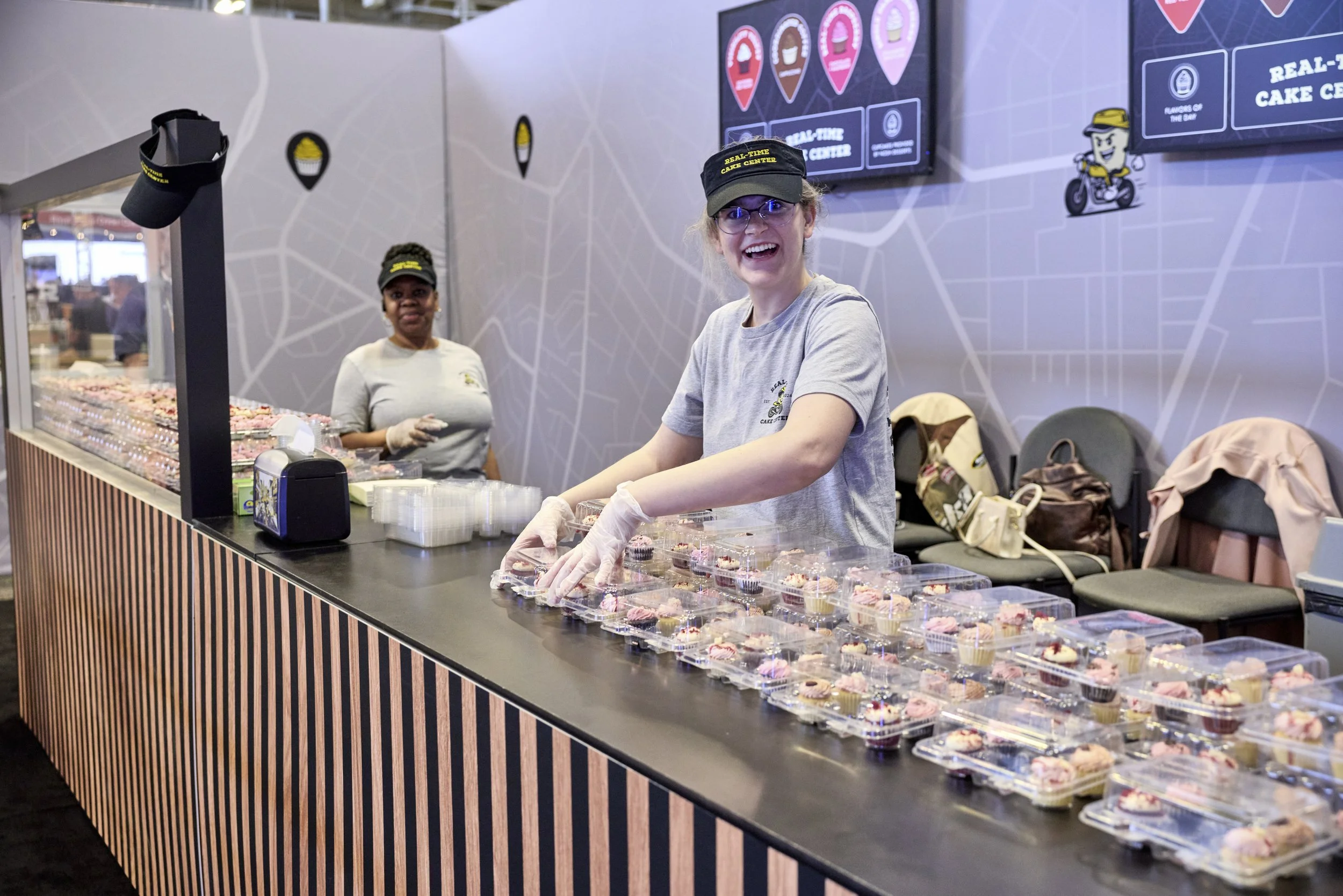 Two women serving cupcakes at a cake center, with a display of cupcakes on the counter and digital menu screens in the background.