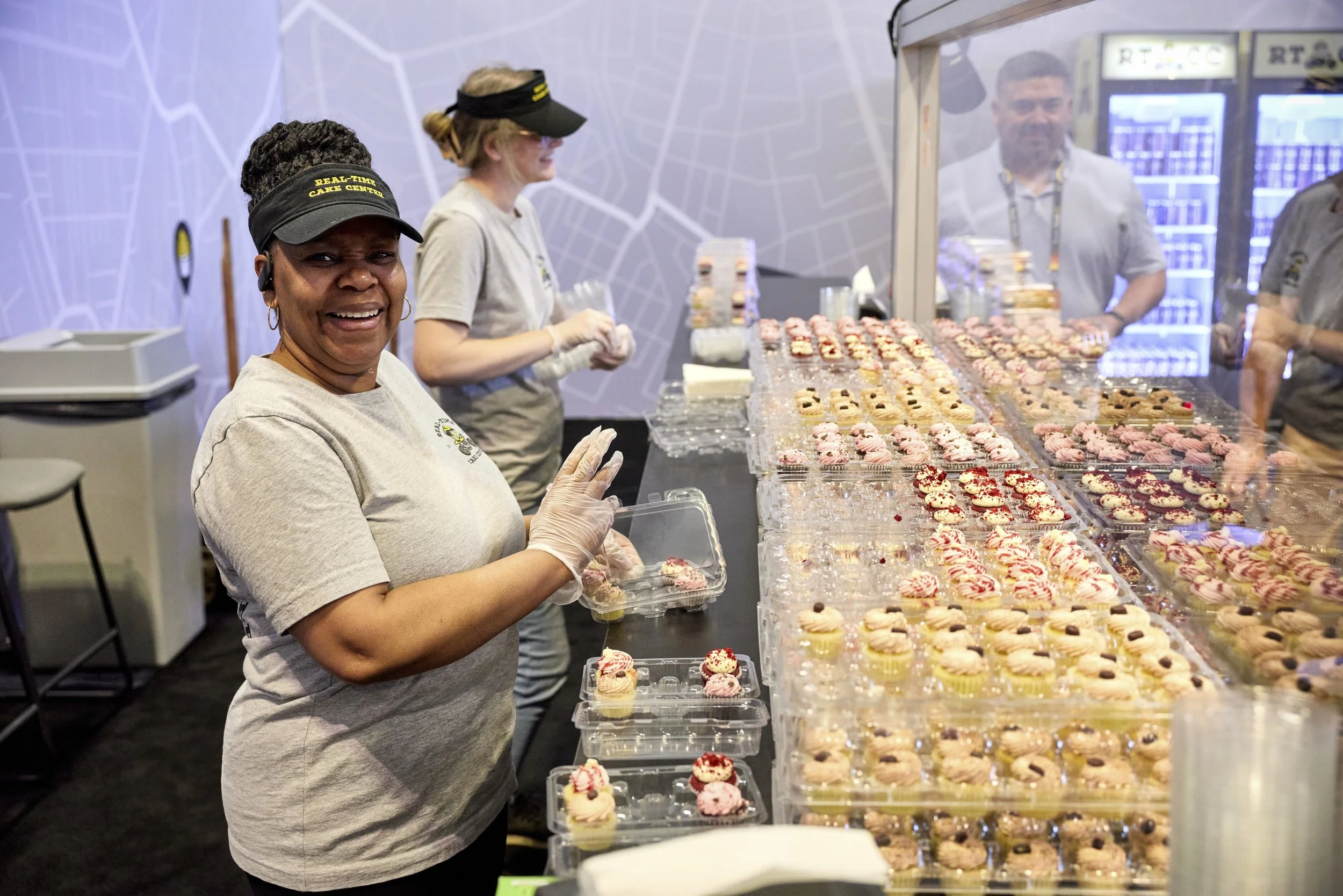 A woman smiling while working at a table of assorted cupcakes with pink, white, and red icing. Other people are seen in the background, some behind a glass partition, in a room with a purple wall featuring a map design.