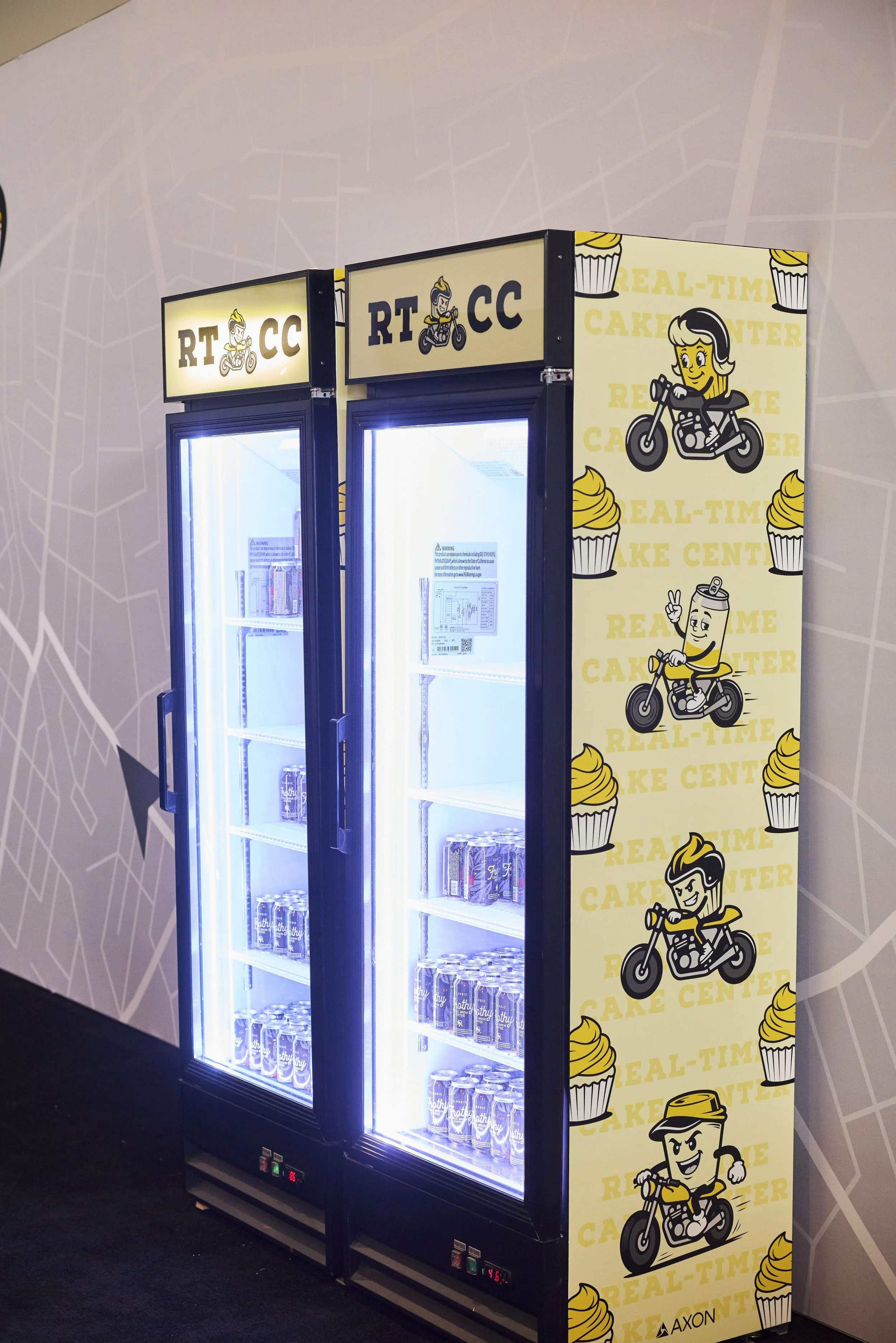 A refrigerated display case with a black frame, illuminated with bright lighting, contains cans and bottles. The side panel features a yellow background with cartoon characters riding motorcycles, cupcake illustrations, and the text "Real-Time Cake C