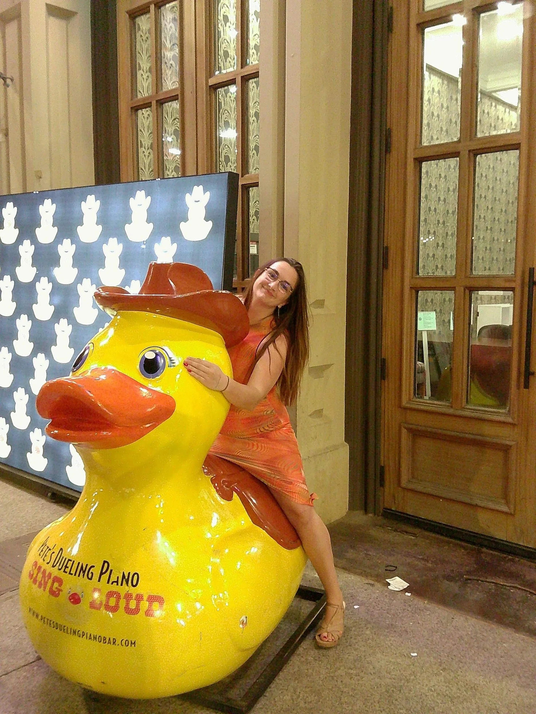 A woman with glasses and long brown hair wearing an orange dress is hugging a large, yellow rubber duck. The duck has a red hat and eyes with a white background. They are inside a room with large windows and wooden frames.