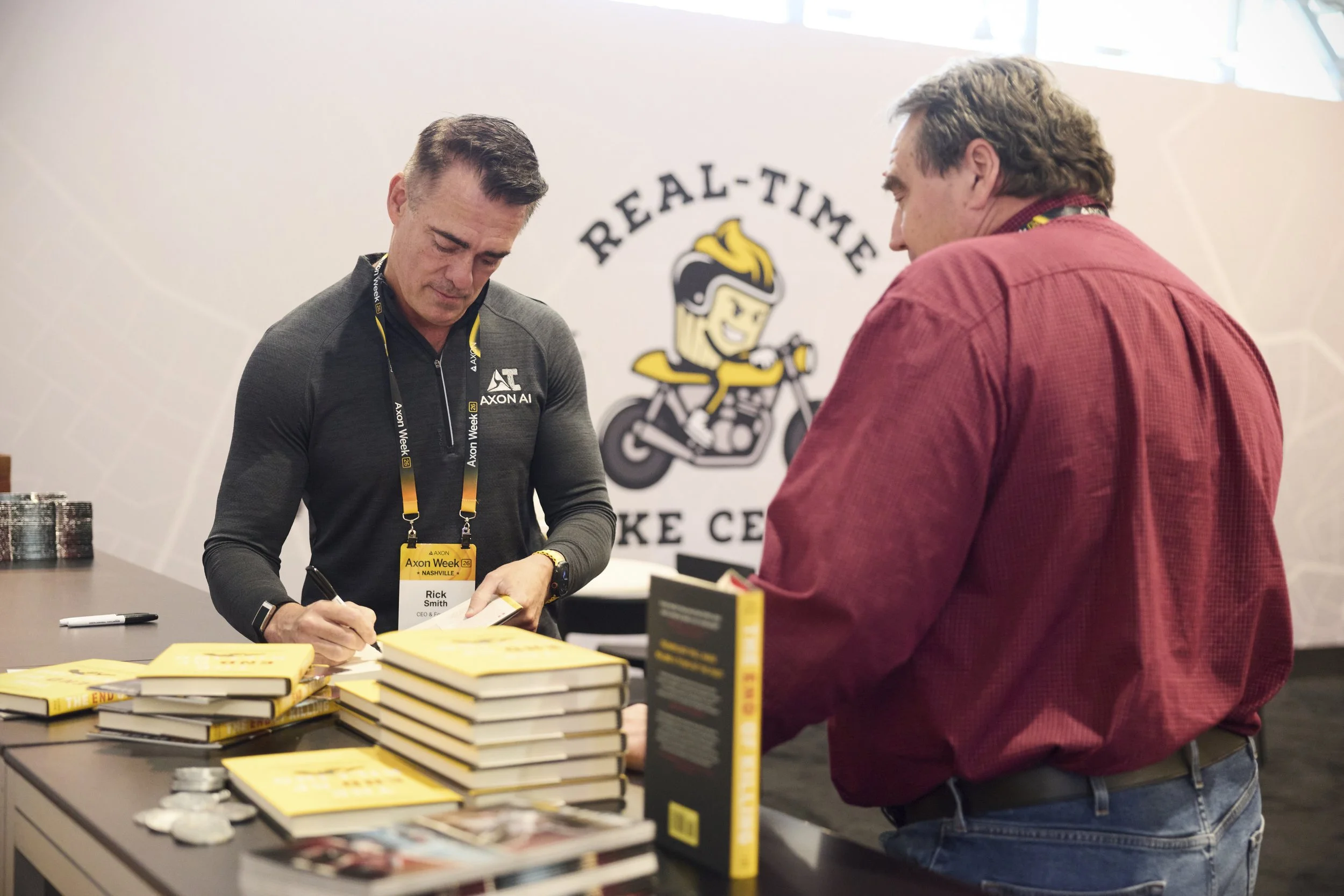 Two men are at a book signing table with stacks of books. The man on the left is signing a book, and the man on the right is facing him. Behind them is a large sign with a stylized cartoon character of a young person with a mohawk riding a bicycle, w