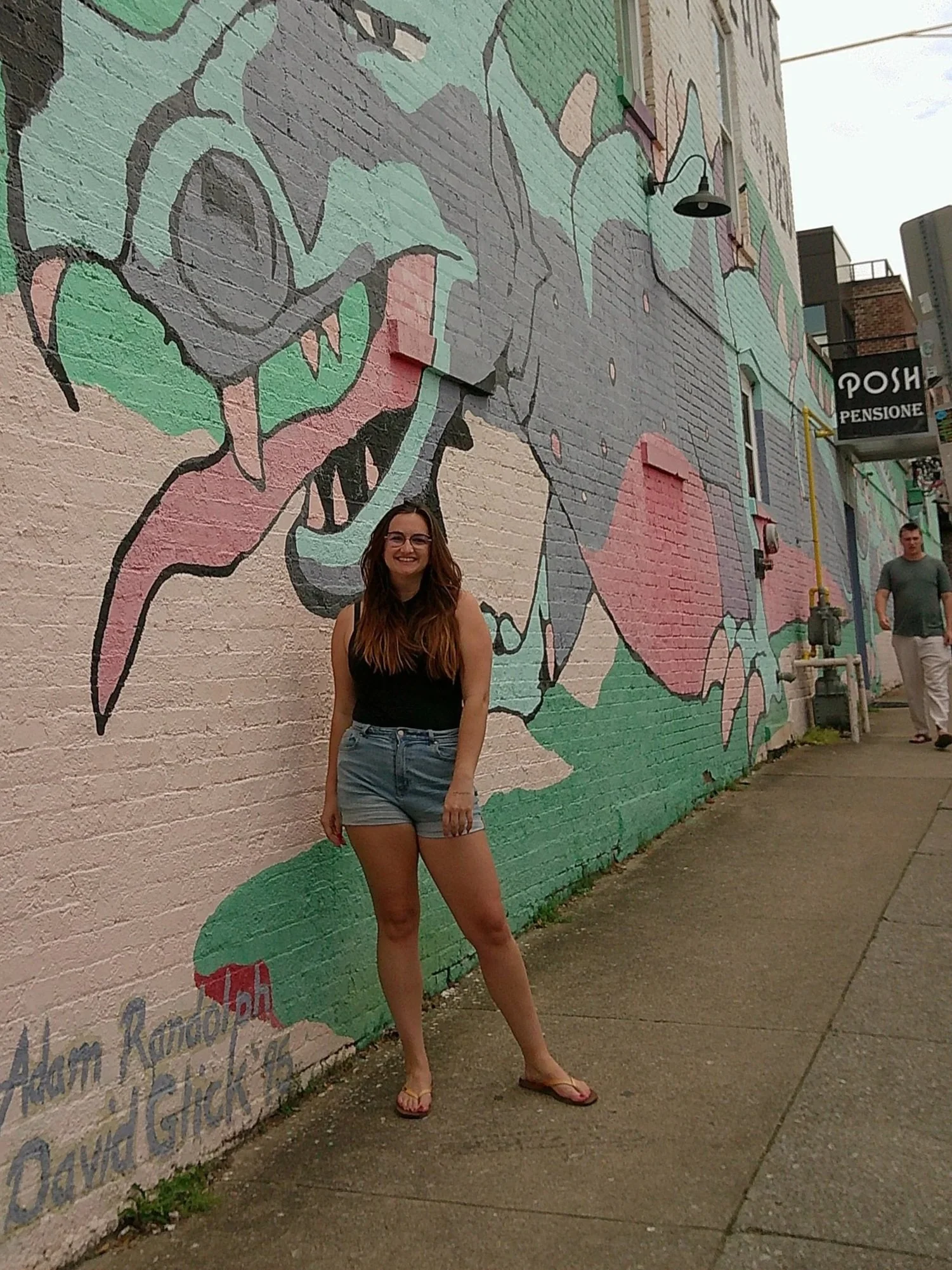 A young woman with long brown hair wearing glasses, a black sleeveless top, and grey shorts, standing outdoors in front of a colorful mural painted on a brick wall. The mural features abstract shapes in pastel colors, including pink, green, and grey.