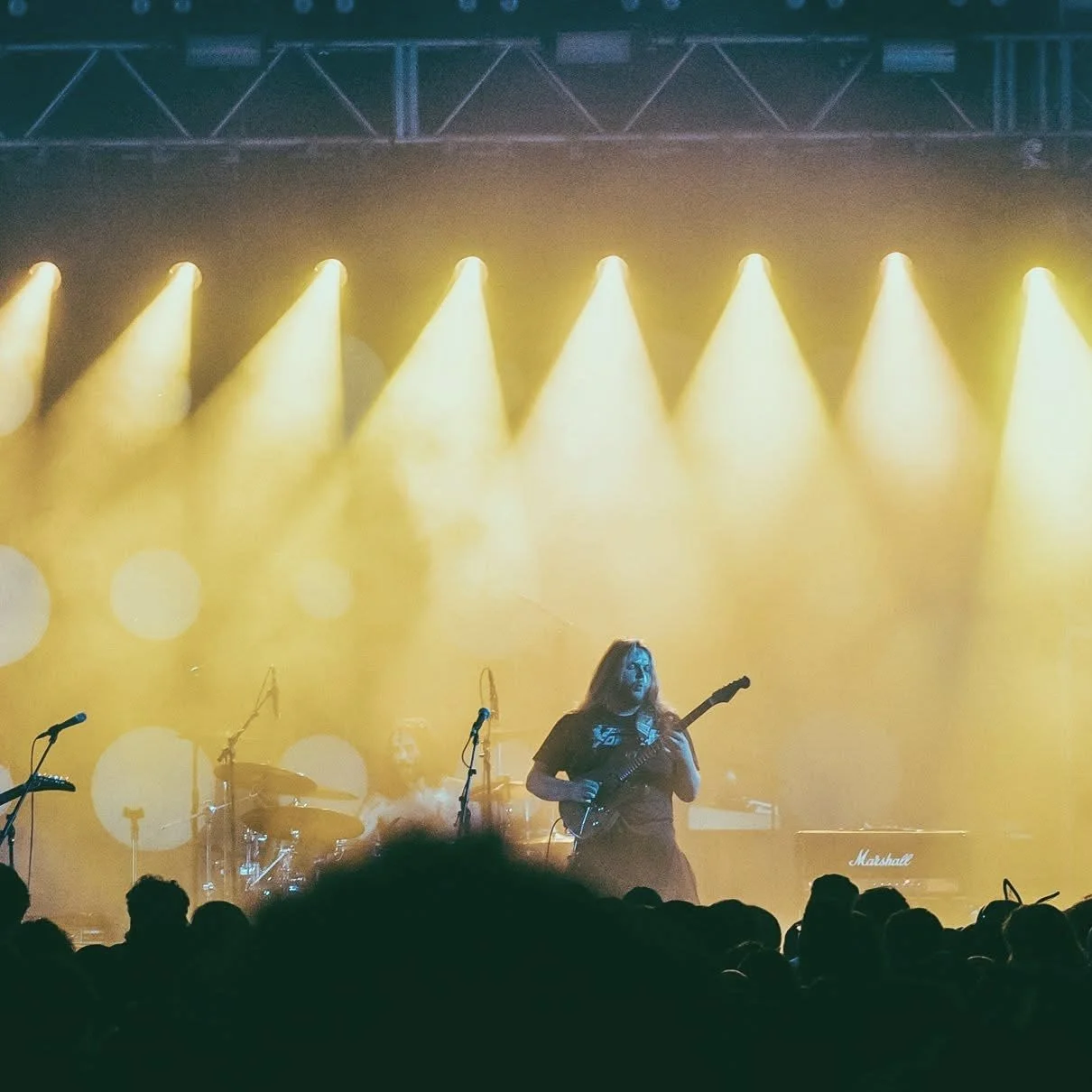 Musician performing on stage with yellow lighting, playing an electric guitar, with a drum set and microphone stands visible, and an audience in the foreground.
