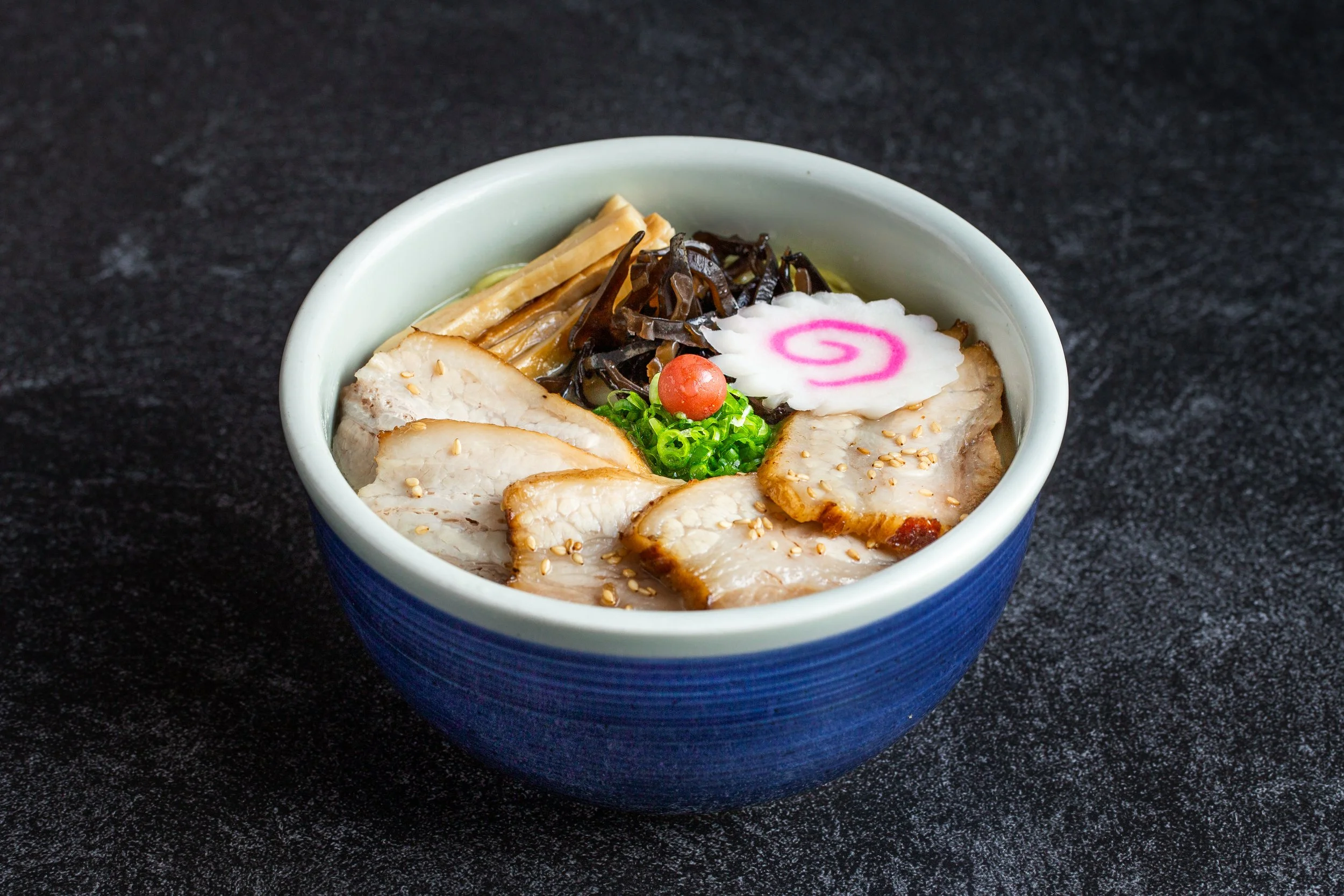 A bowl of ramen with 5 pieces of cha-siu along with naruto, green onions, umeboshi, etc.