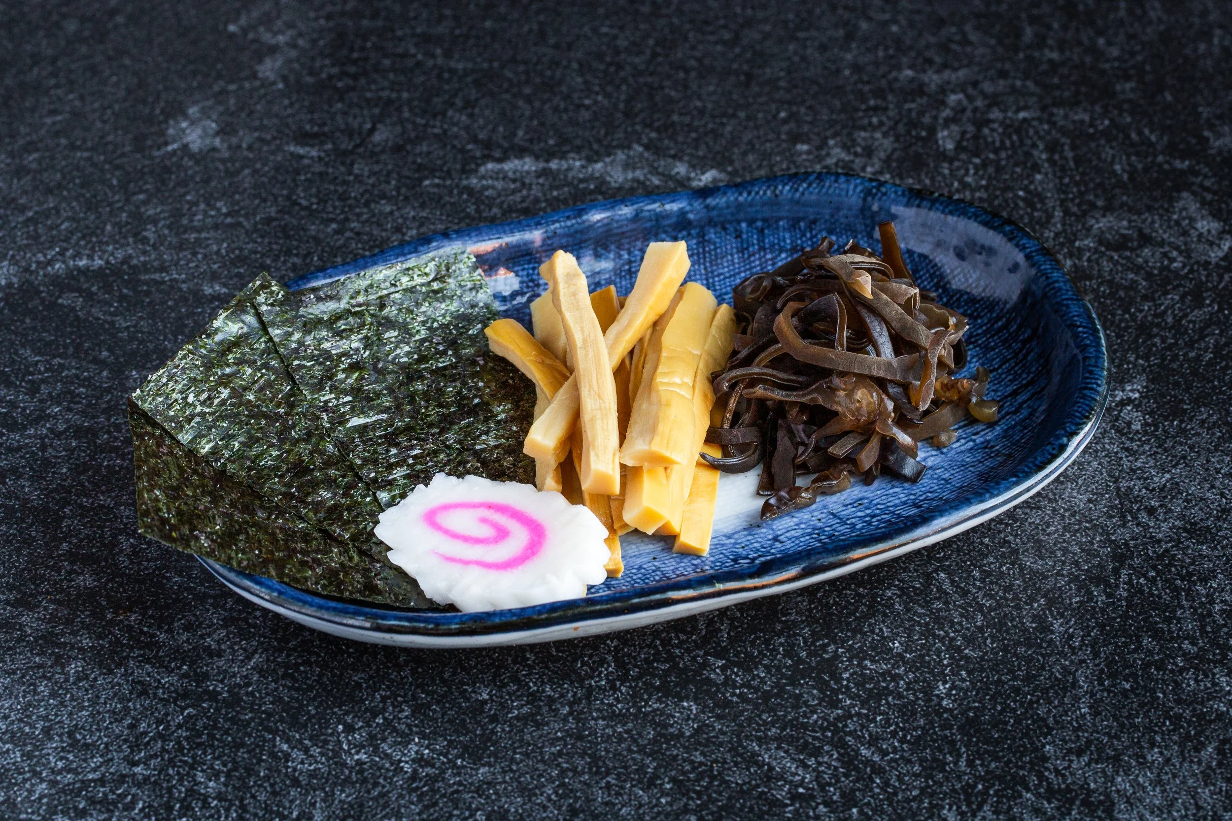 Dry seaweed, bamboo shoots, kikurage with a slice of naruto on a blue plate