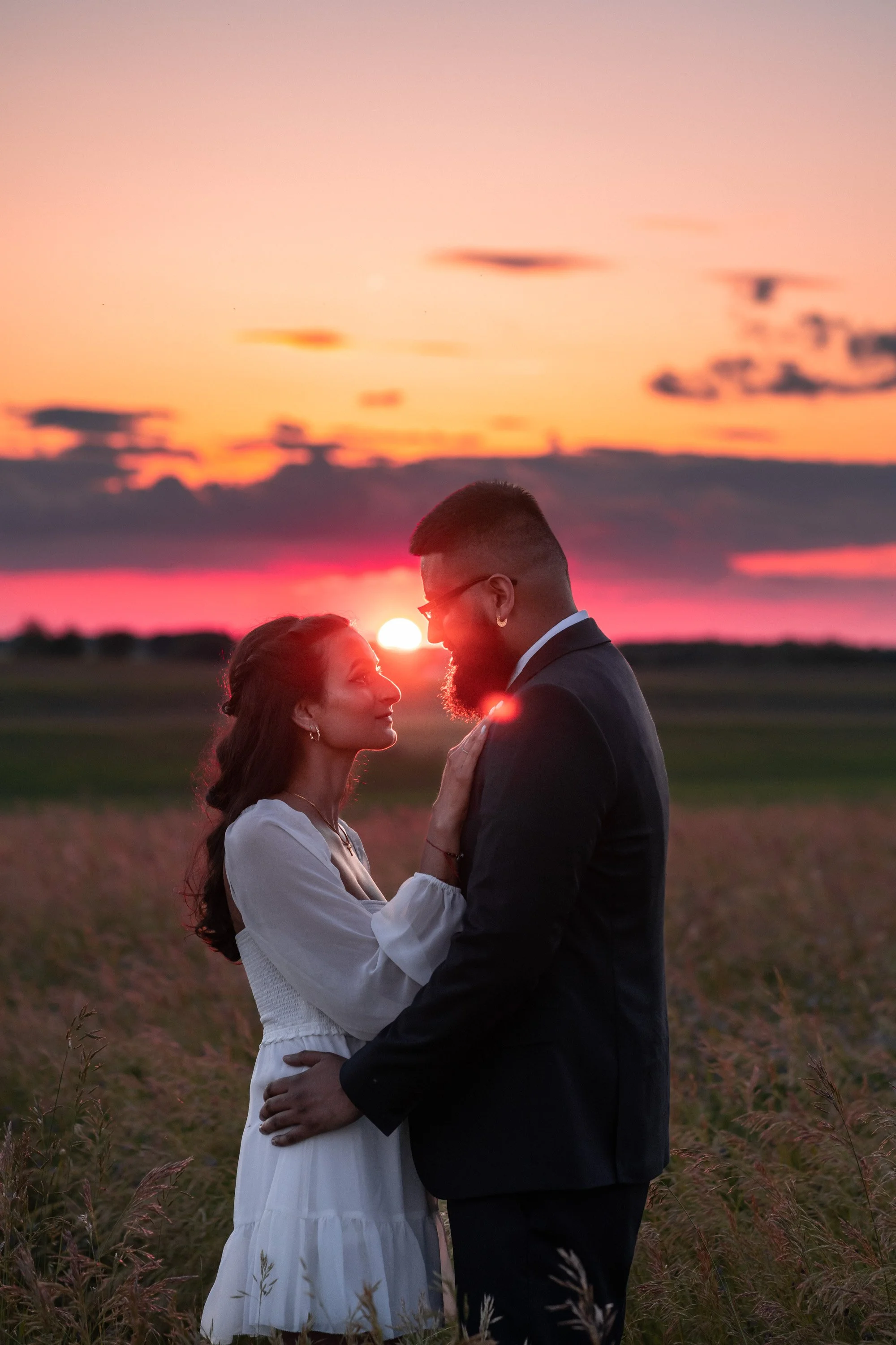 A couple dressed formally standing close in a field during sunset, gazing into each other's eyes with the sun setting on the horizon behind them.