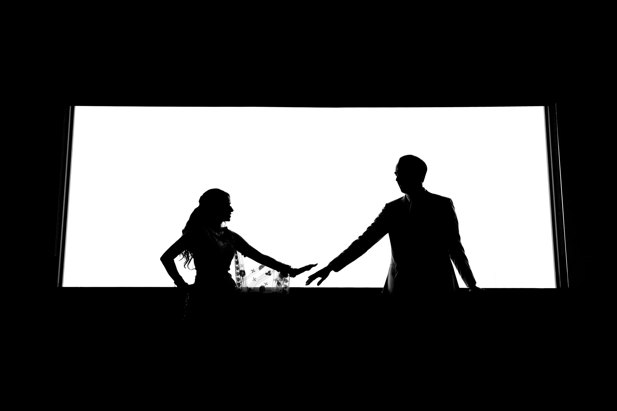 Silhouette of a man and woman holding hands in front of a bright rectangular background, with the woman wearing a flowing dress and the man in a suit.