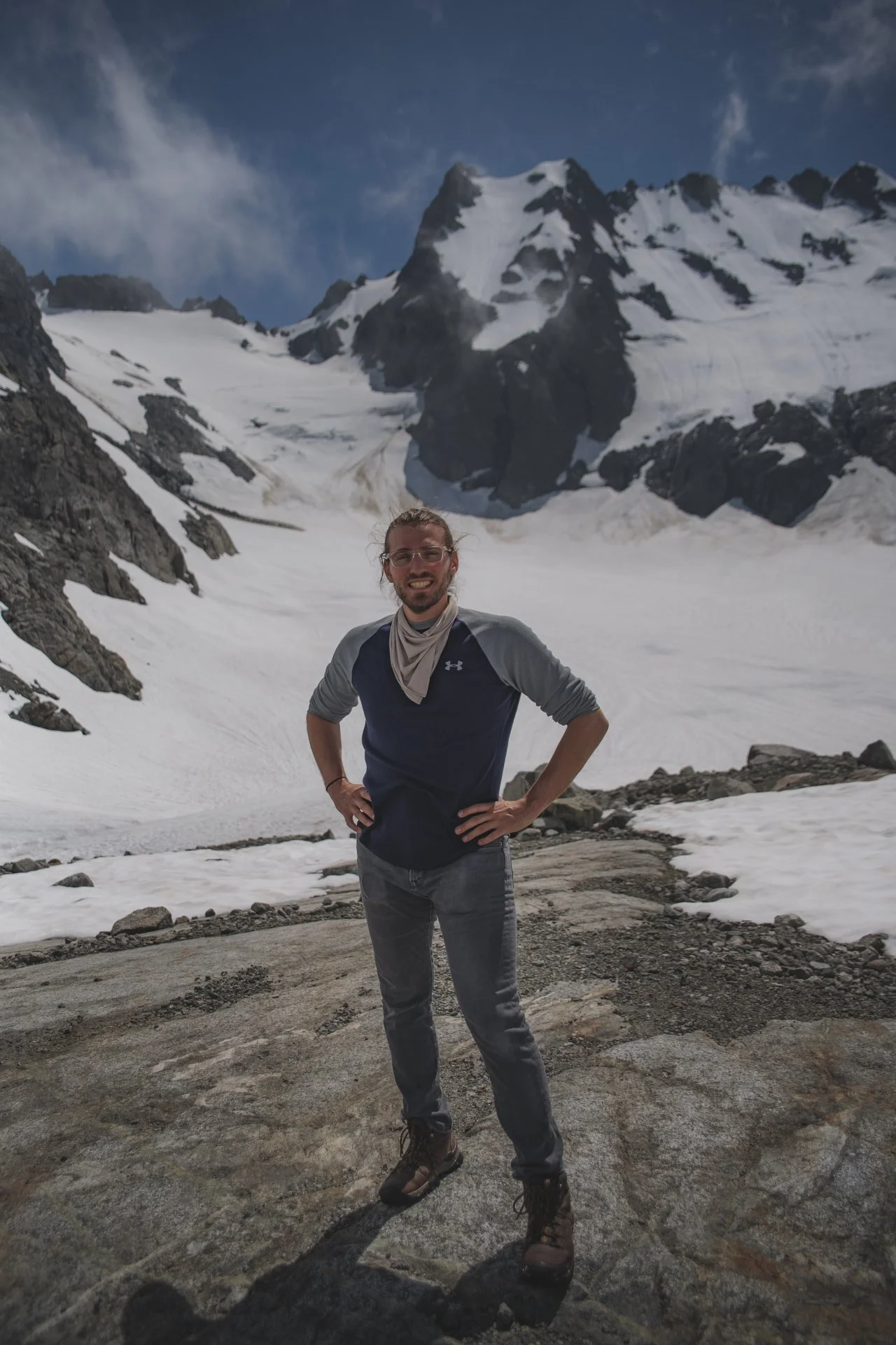 A man standing with his hands on his hips in a snowy mountainous landscape, with snow-covered peaks and a partly cloudy sky in the background.