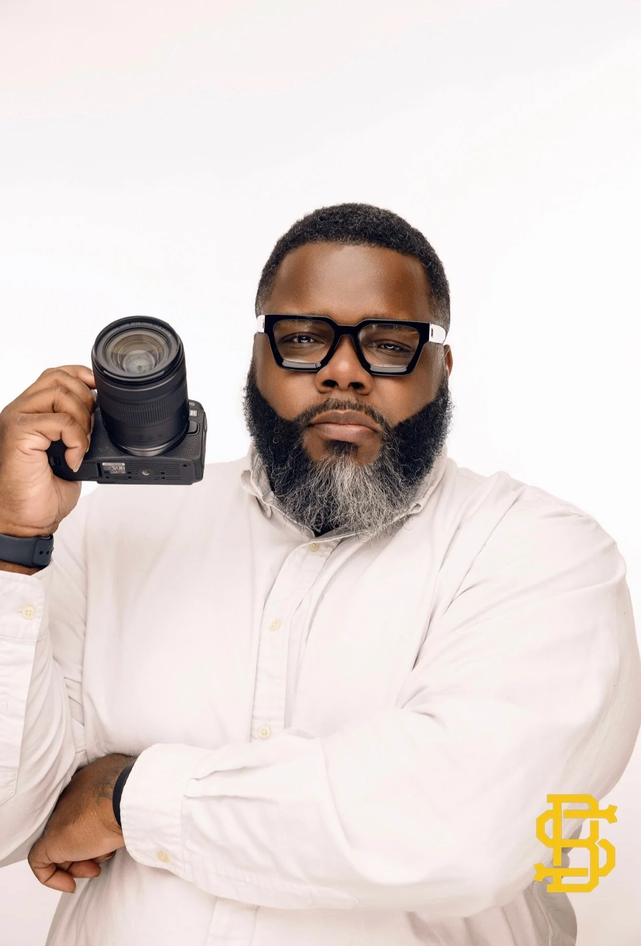 Portrait of a man with a beard and glasses wearing a white shirt, holding a camera, with crossed arms in front of a plain white background. A yellow logo is in the bottom right corner.