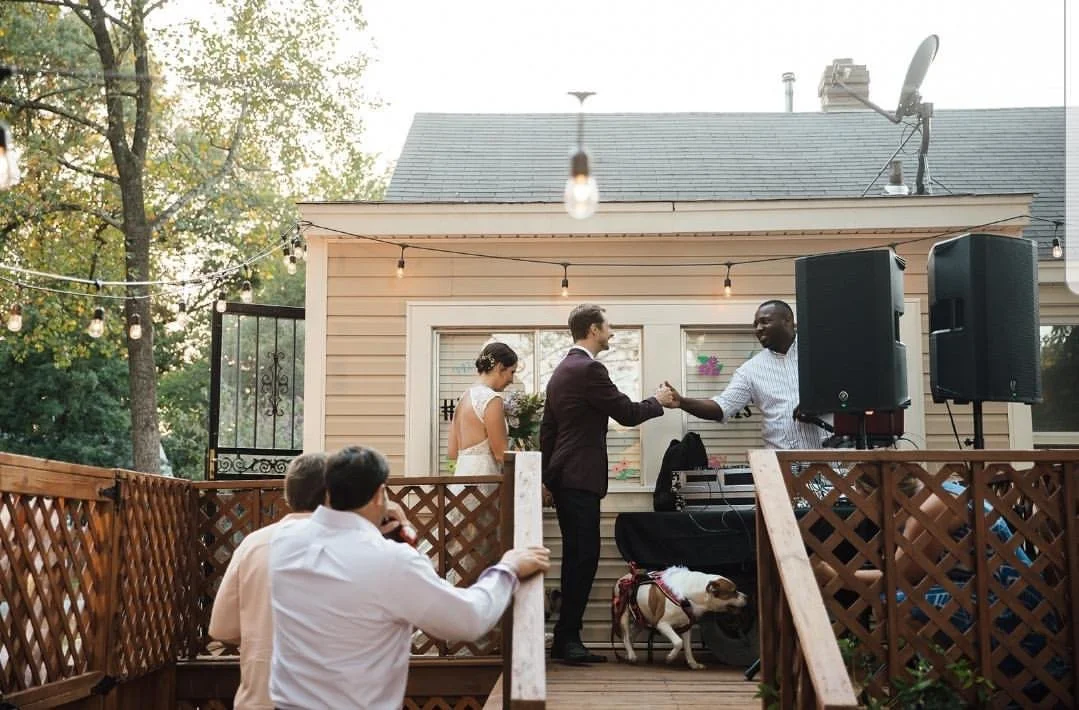 A wedding reception on an outdoor deck with a bride, groom, and officiant, with guests seated nearby and string lights above. Wedding DJ
