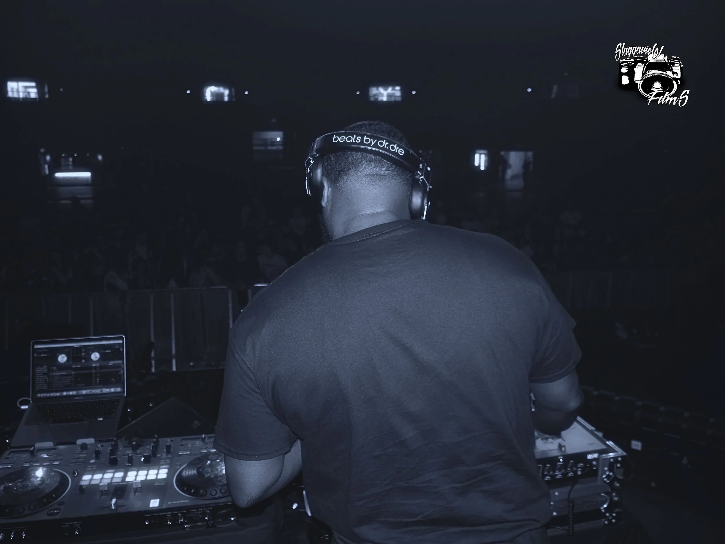 A DJ wearing headphones with text 'beats by dr. dre' on them, working at a DJ booth with turntables and a laptop in a dark setting.