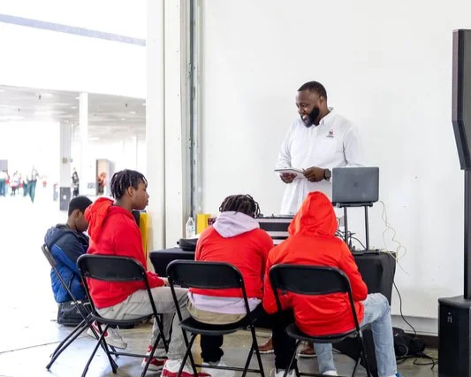 A man standing in front of four kids sitting at a table, teaching or presenting with a laptop, in a bright indoor space. Mentoring the kids